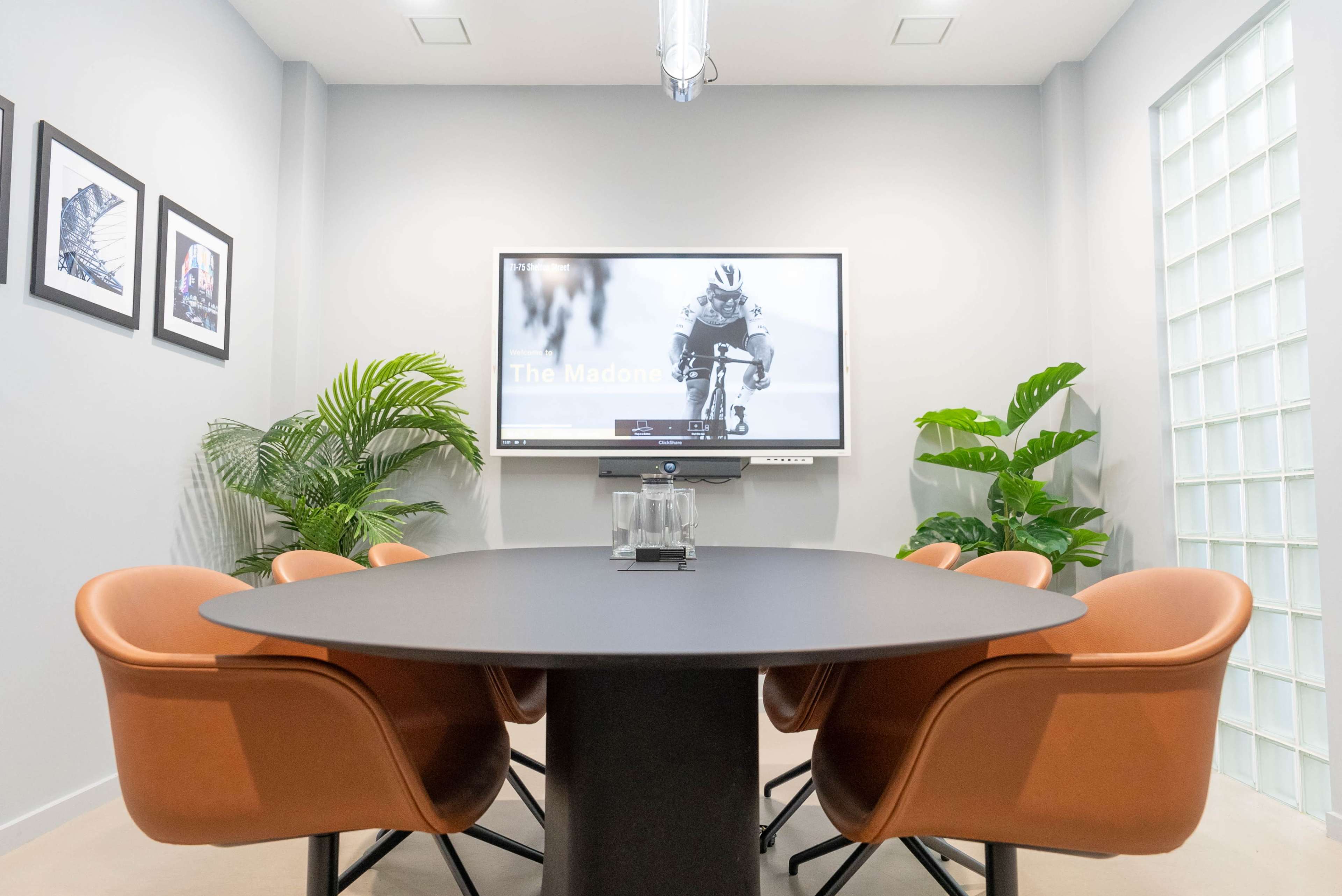 A conference room features a round table surrounded by brown chairs, with a wall-mounted screen displaying an image of a cyclist and potted plants in the background.