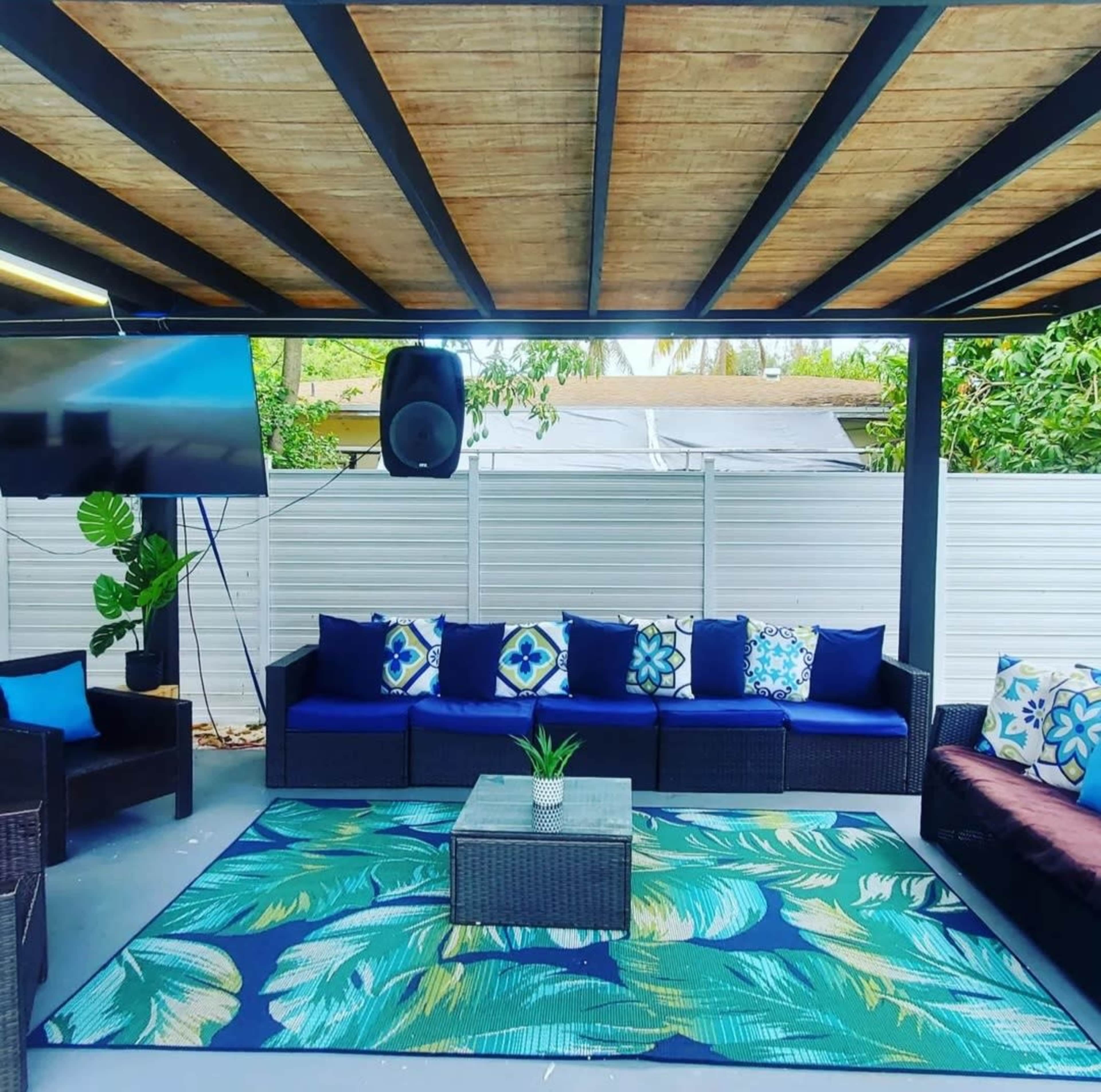 A covered outdoor seating area with a sectional sofa, decorative cushions, a coffee table, and a large television mounted on the wall.