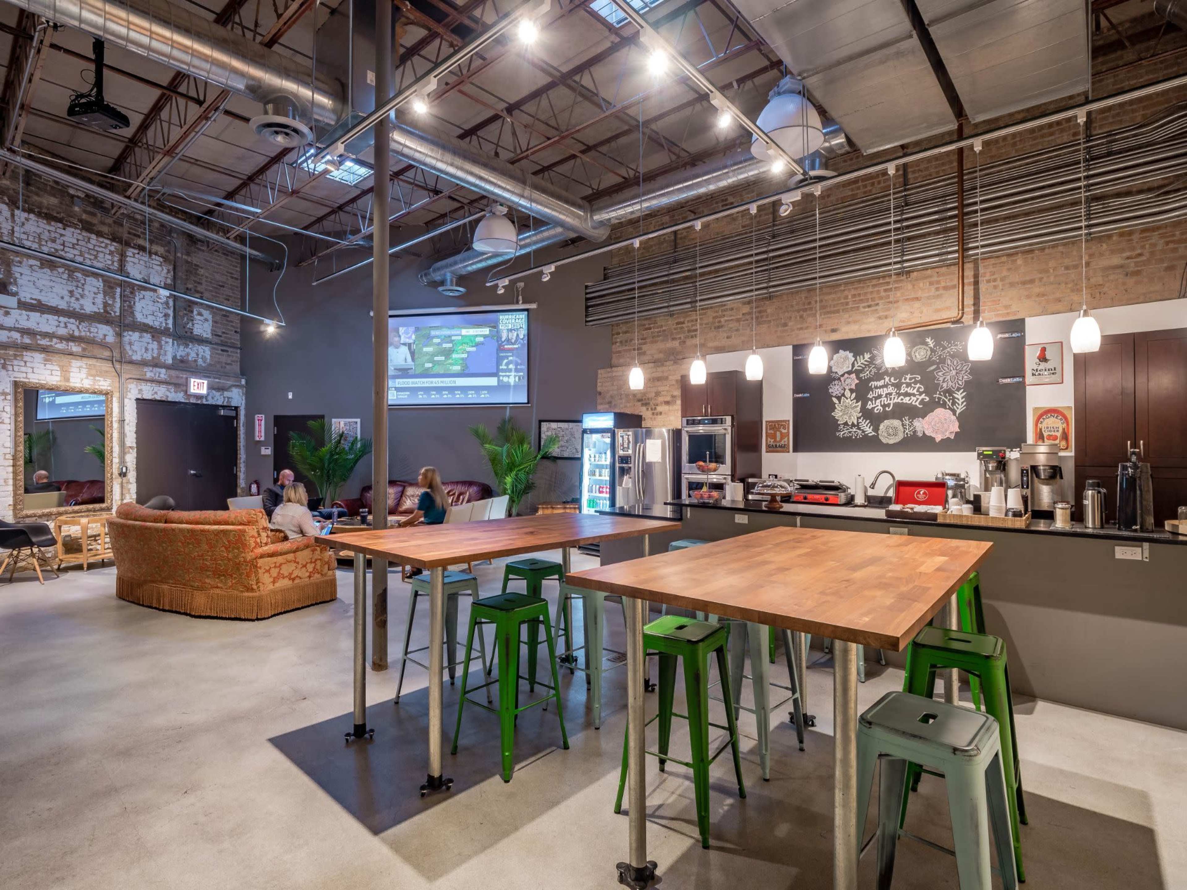 A spacious cafe features industrial decor with high tables, metal stools, a coffee machine, and a large screen displaying content on a brick wall.