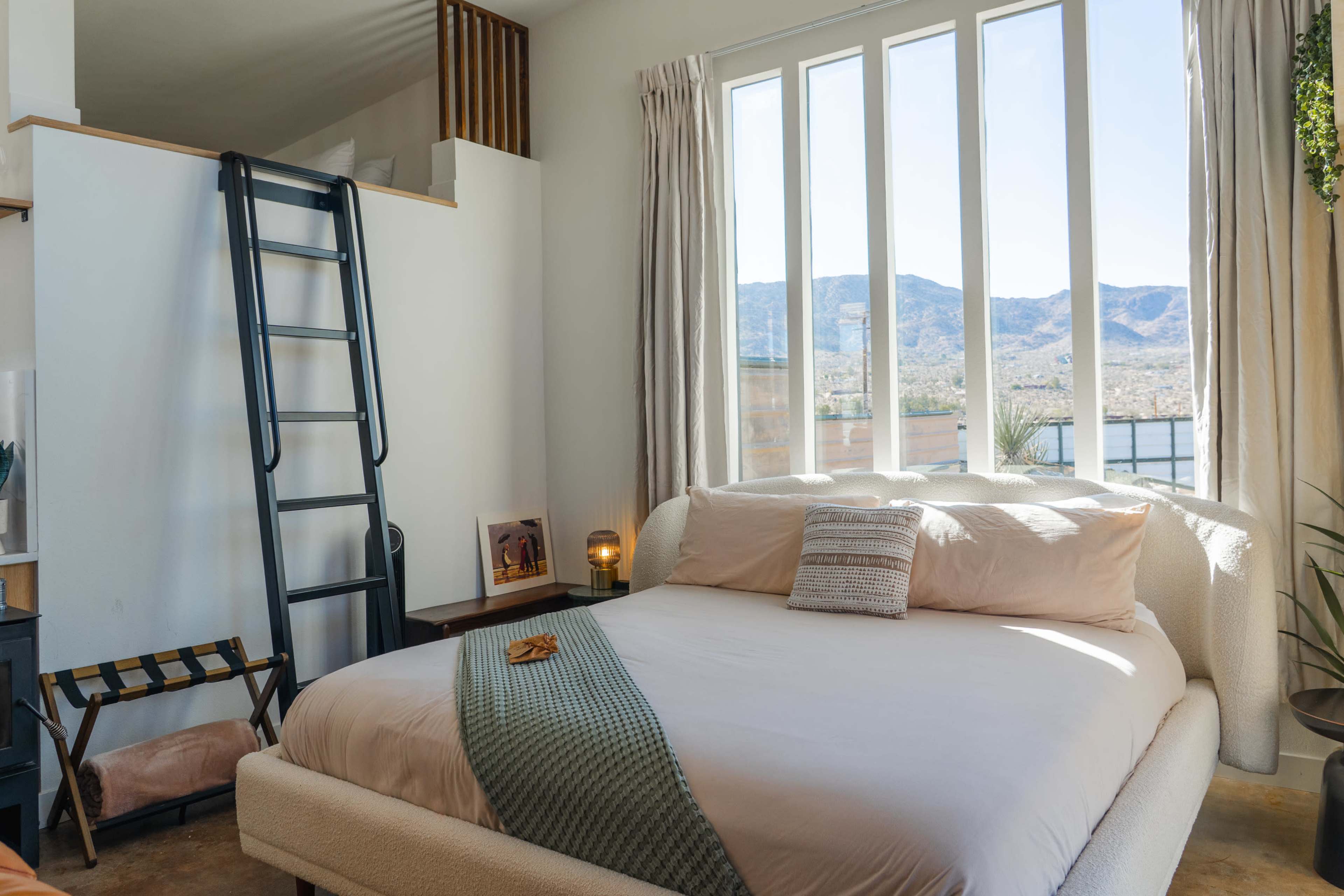 A neatly arranged bedroom features a large bed with light-colored bedding, a tall window offering mountain views, and a ladder leading to a lofted area.