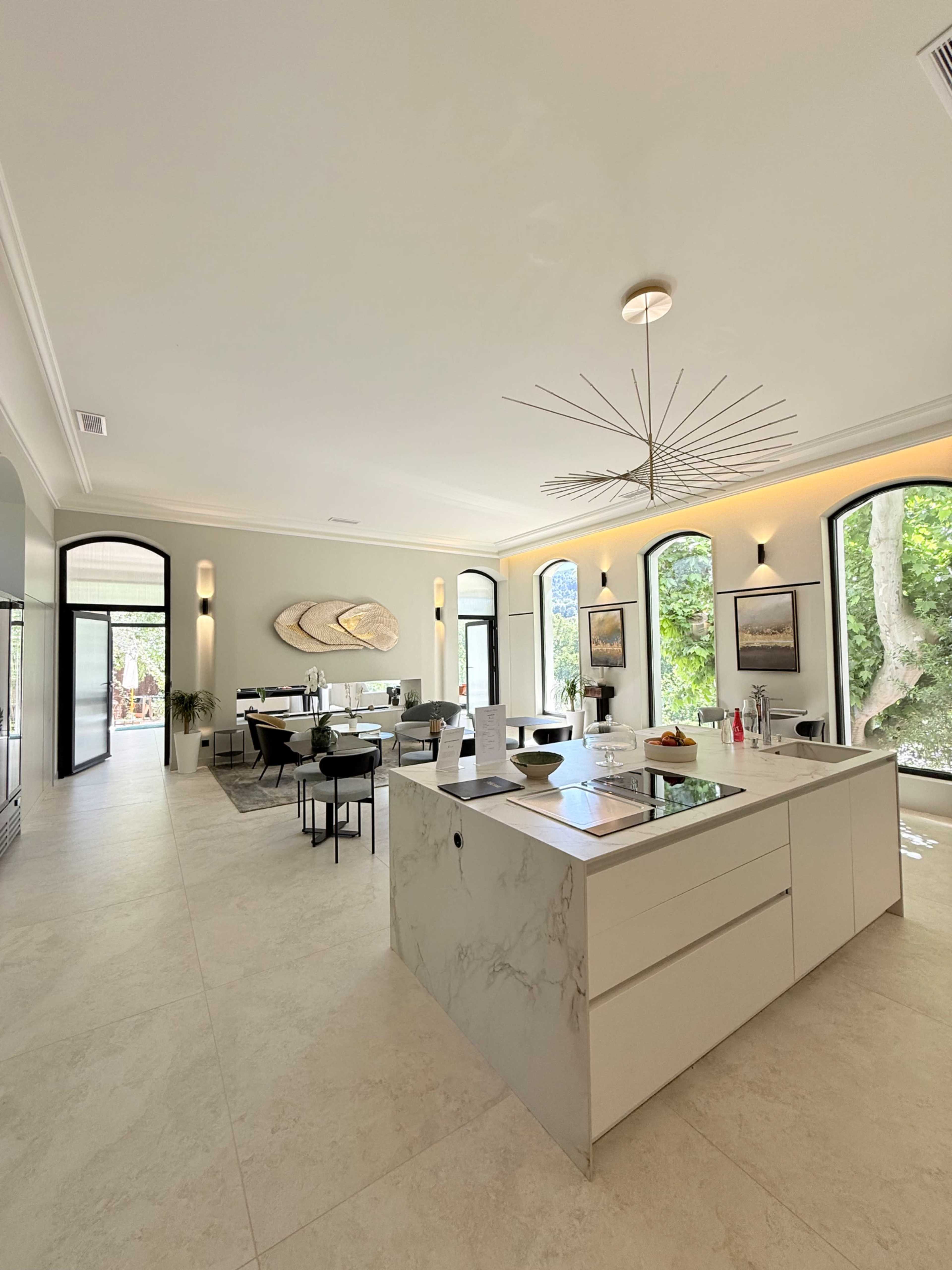 The image shows a modern, open-concept kitchen and dining area with large arched windows, a central island, and minimalist furnishings.