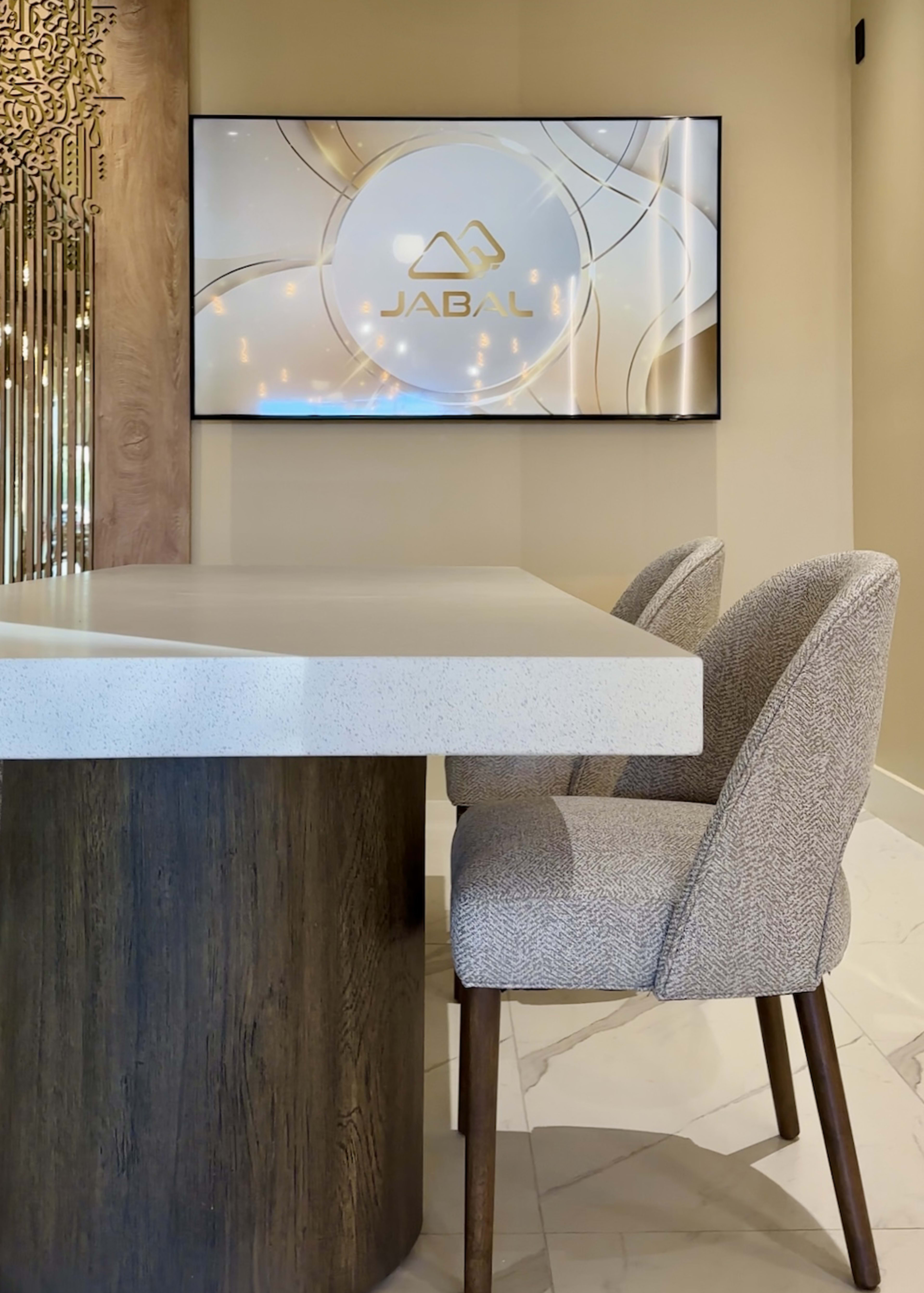 A modern reception area features a sleek table with a textured chair and a wall-mounted screen displaying the logo for "JABAL."