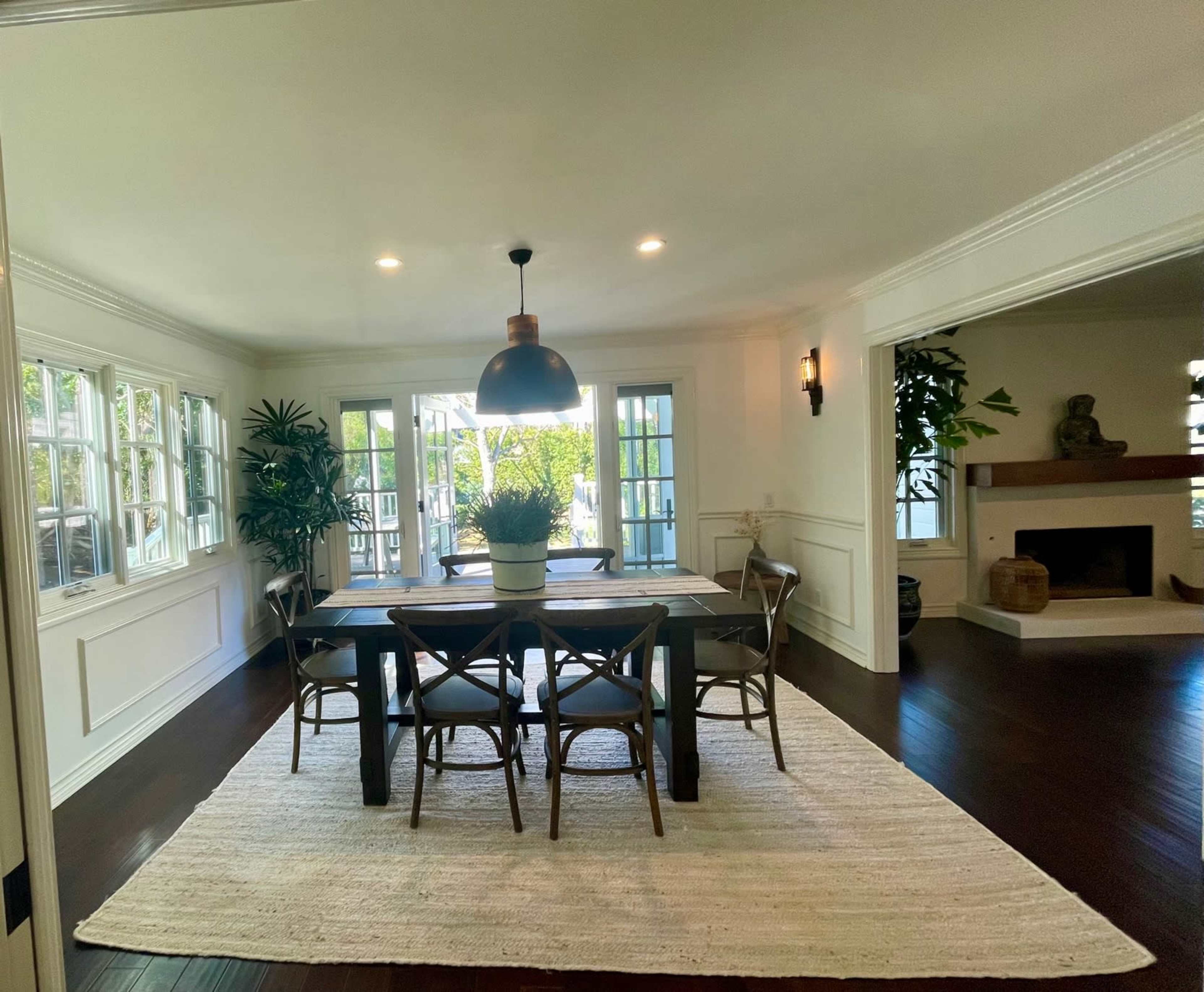 A dining room features a large wooden table surrounded by six chairs, with natural light streaming through multiple windows and a fireplace visible in the adjacent space.