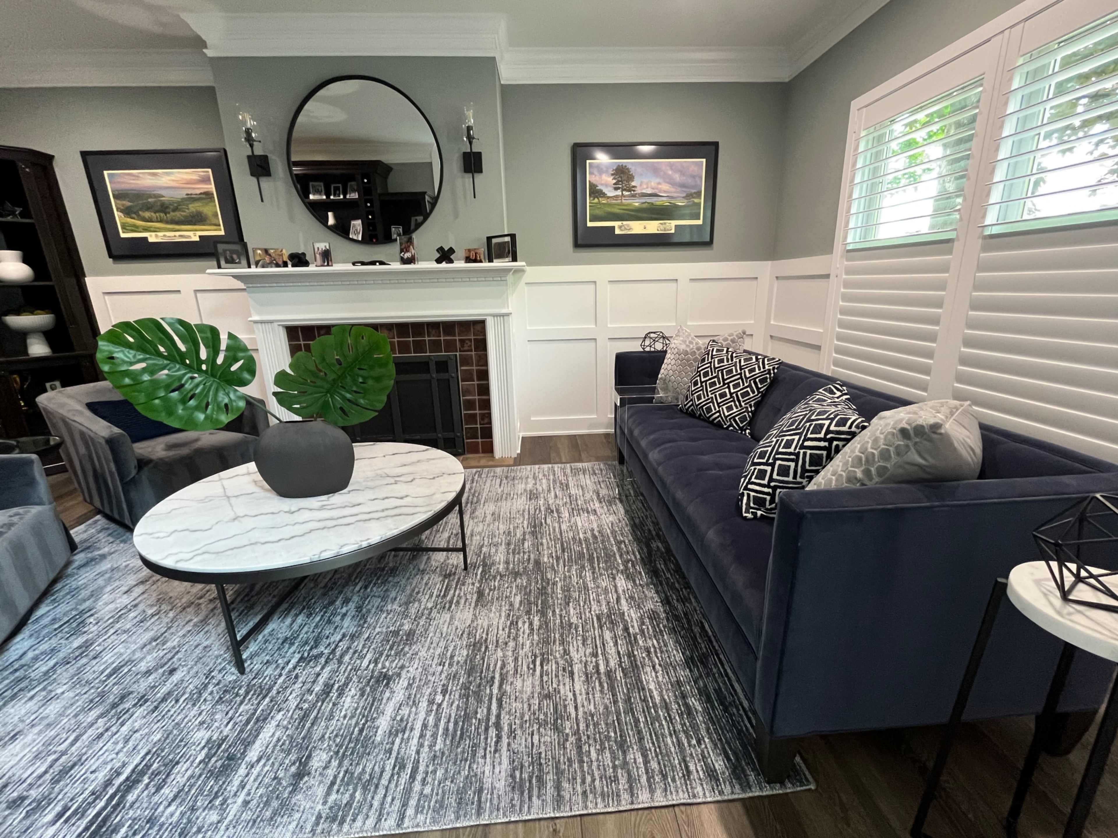 The image shows a living room featuring a dark blue sofa, a round marble coffee table, and framed landscape artwork above a white mantelpiece.