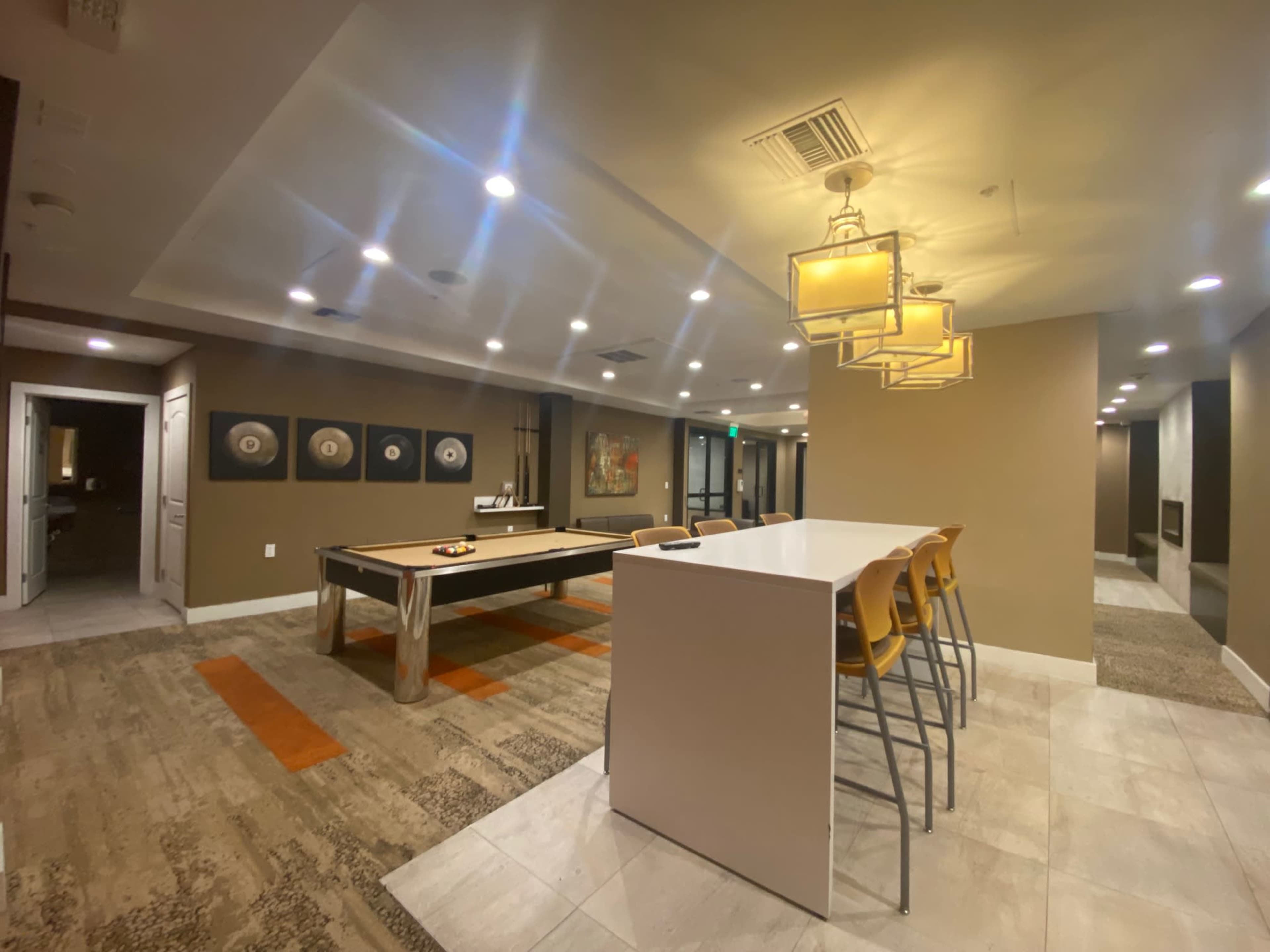 The image shows a modern common area featuring a game table, a white countertop with yellow stools, and a series of pendant lights.
