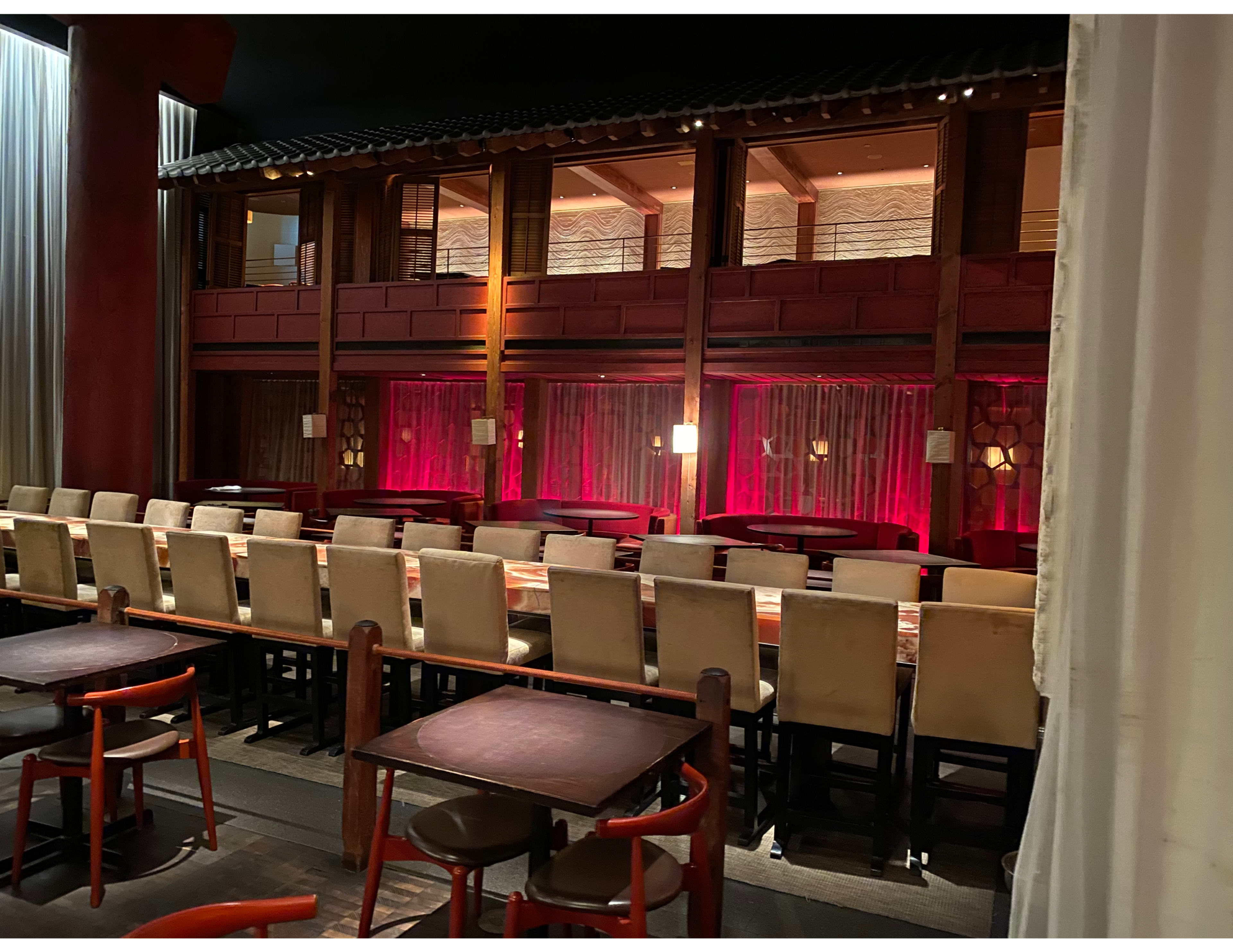 A restaurant interior with a long bar area and elevated seating in a two-level design, featuring red lighting and wooden accents.