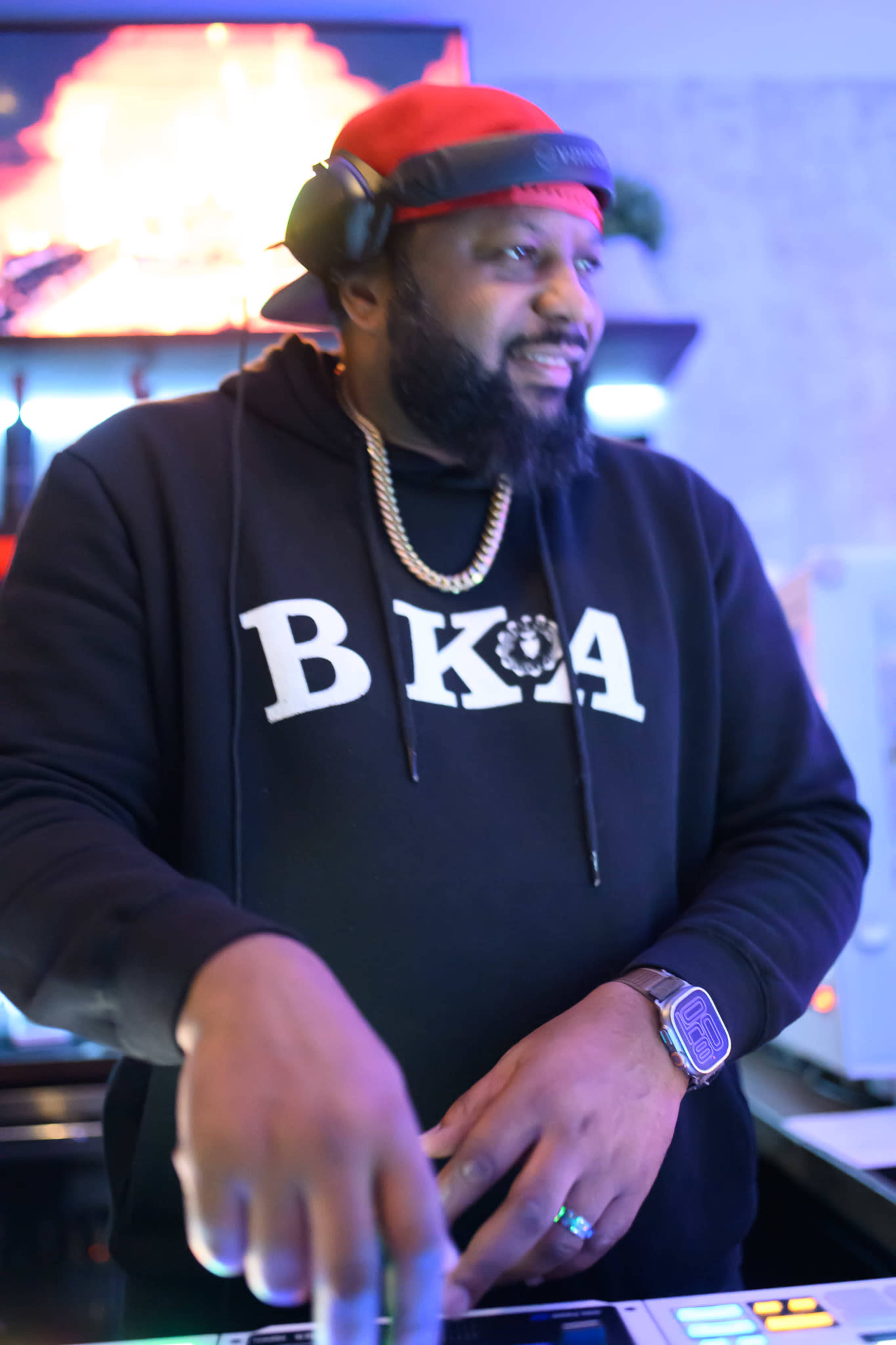 A DJ wearing a black hoodie with "BKA" printed on it performs at a vibrant event with colorful lighting in the background.