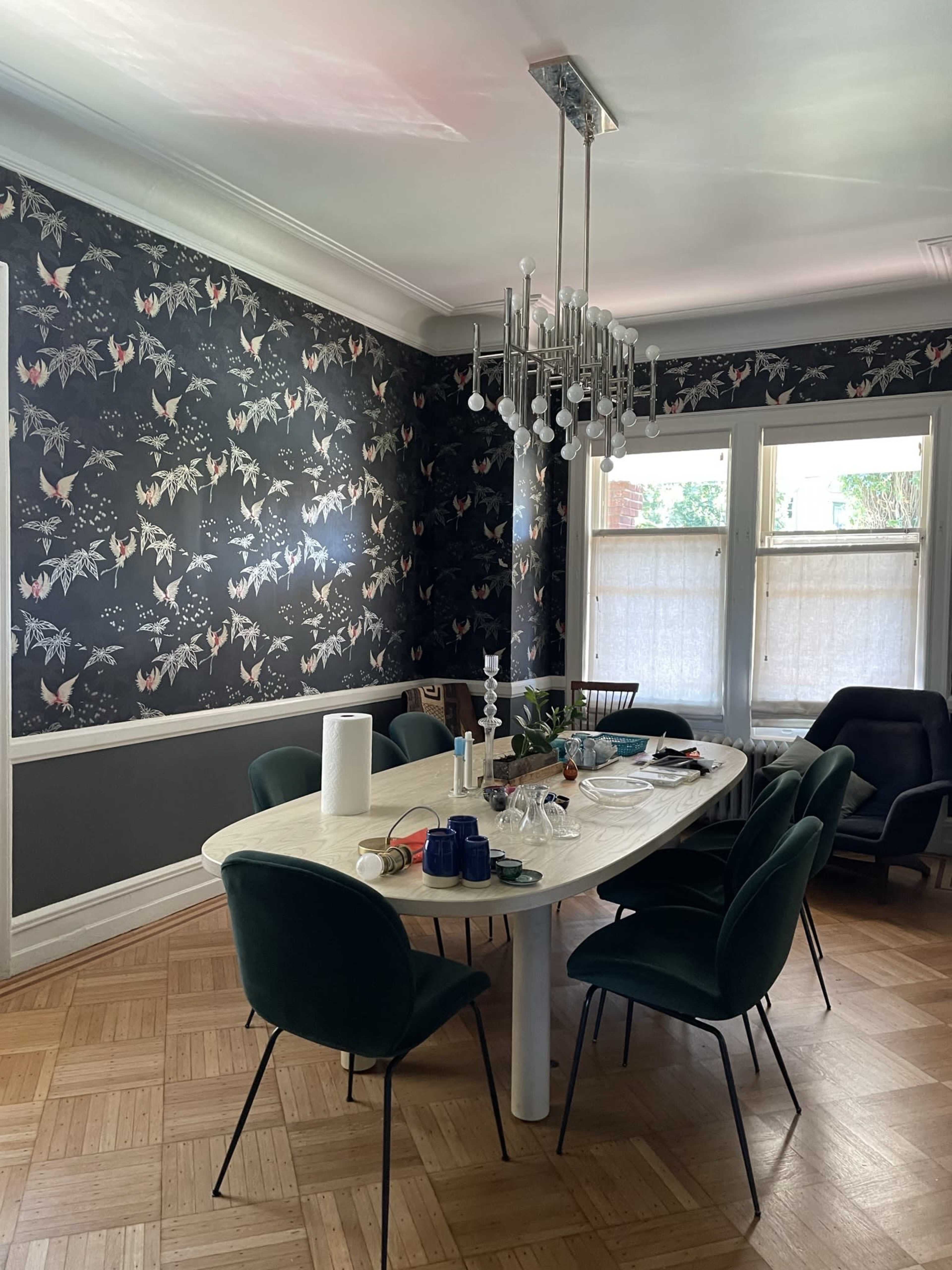 A dining room features a large oval table surrounded by green chairs, with floral wallpaper and a modern chandelier overhead.