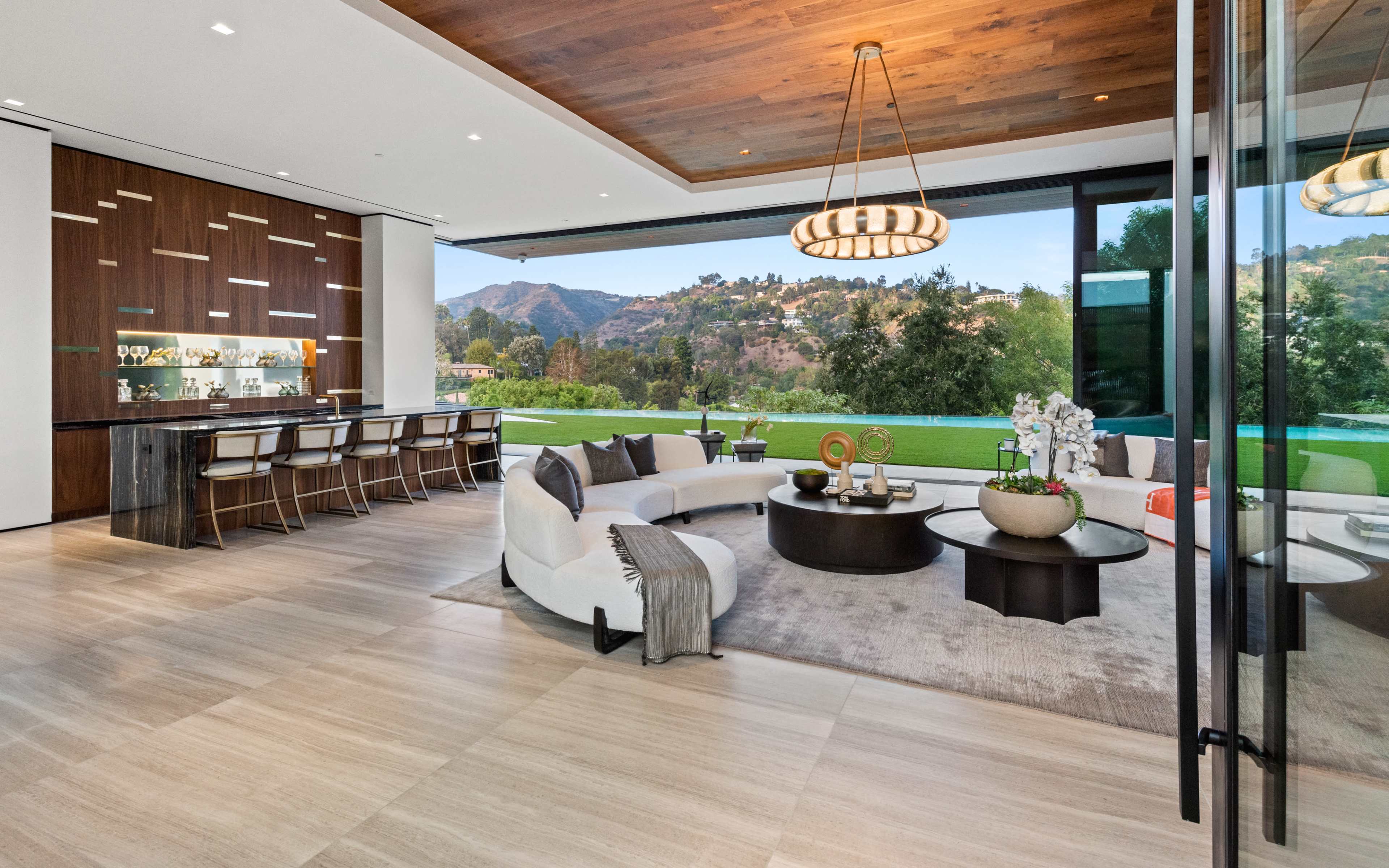 The modern living room features a large open space with a circular coffee table, sectional sofa, and a view of the outdoor landscape through expansive glass windows.