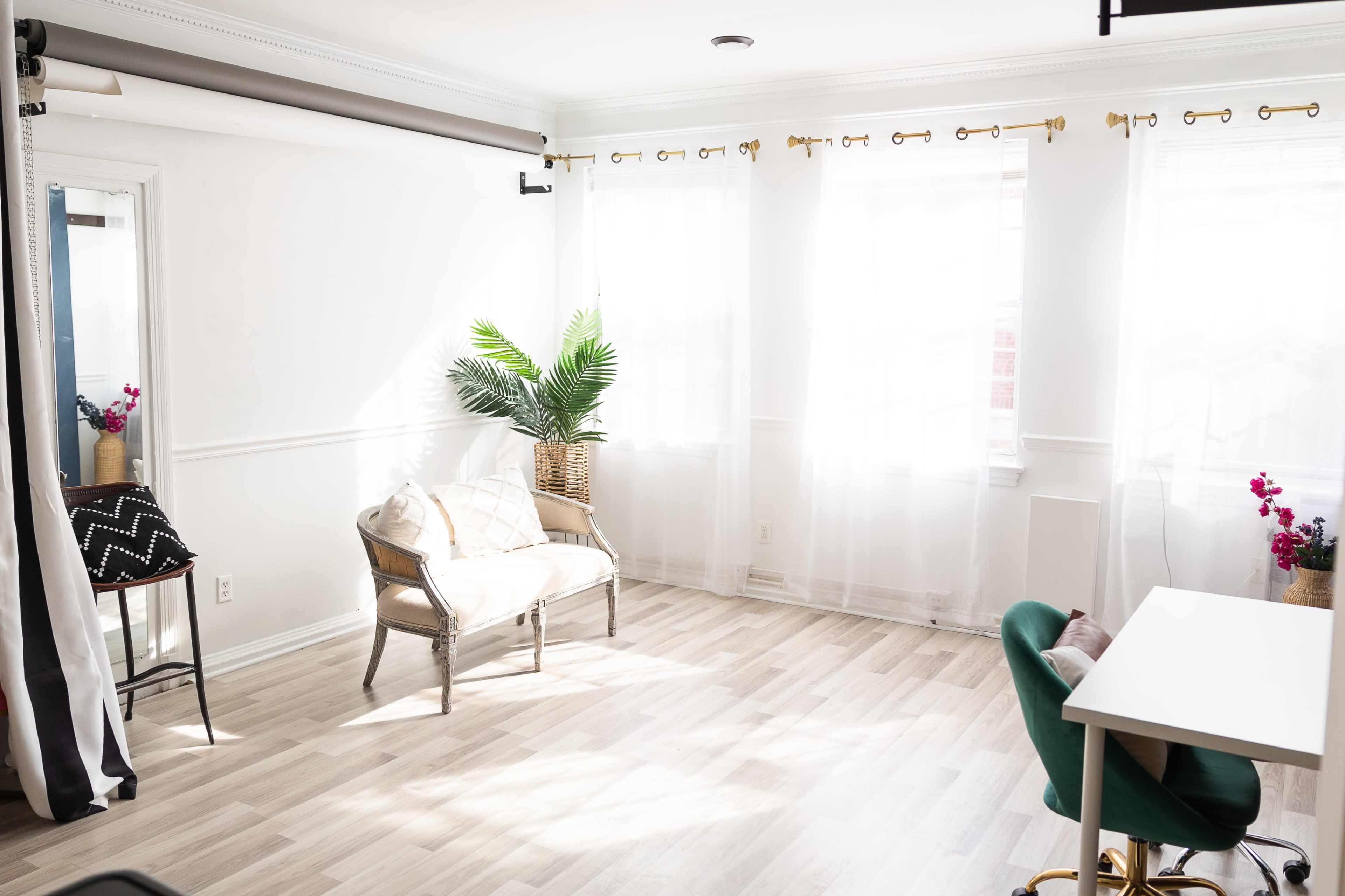 A bright room with large windows, light wood flooring, and simple furniture, including a chair and a desk.