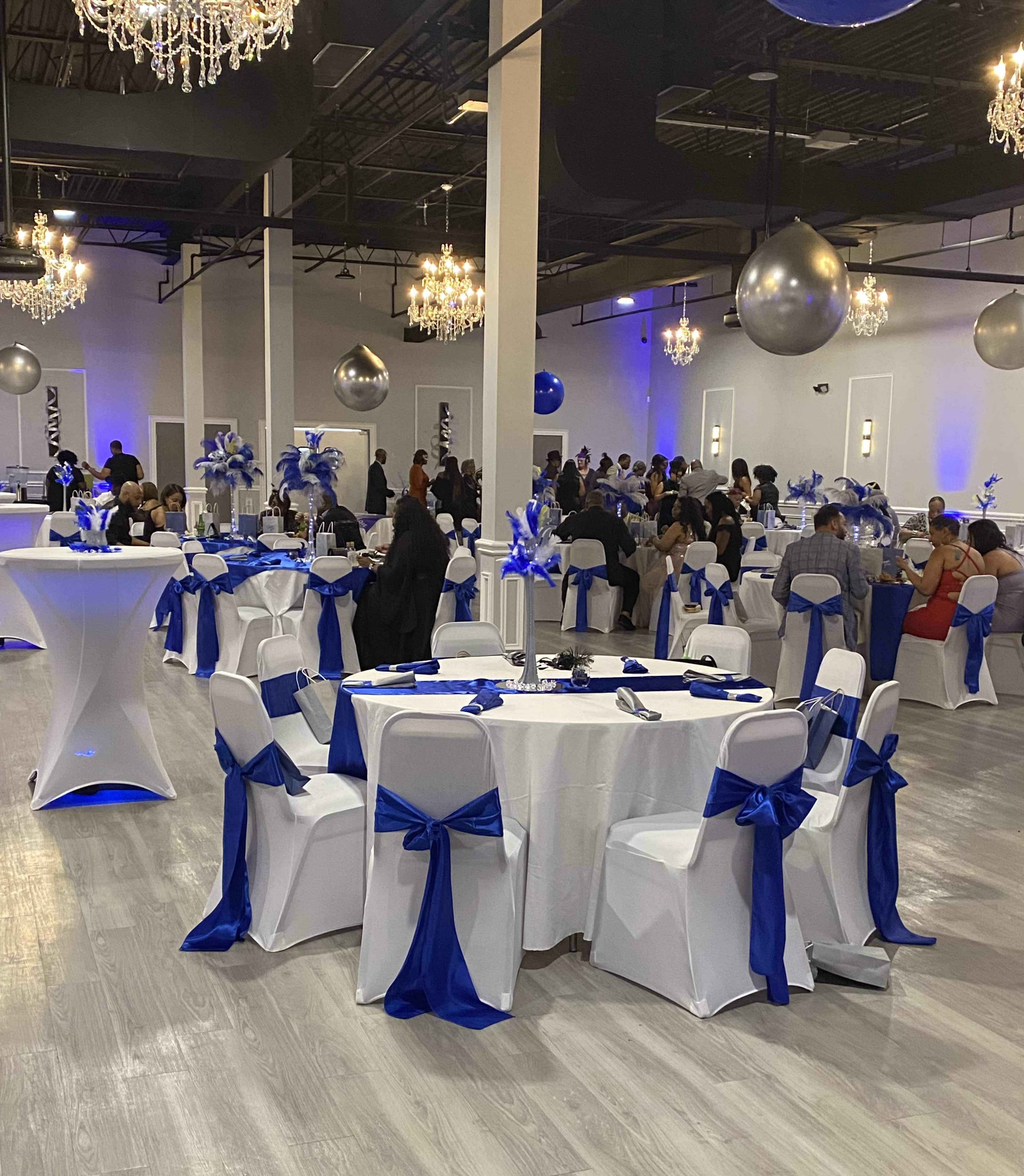 A spacious event venue is set up with white tables adorned with blue satin bows, featuring chandeliers and large silver decor elements hanging from the ceiling, while guests mingle throughout the room.