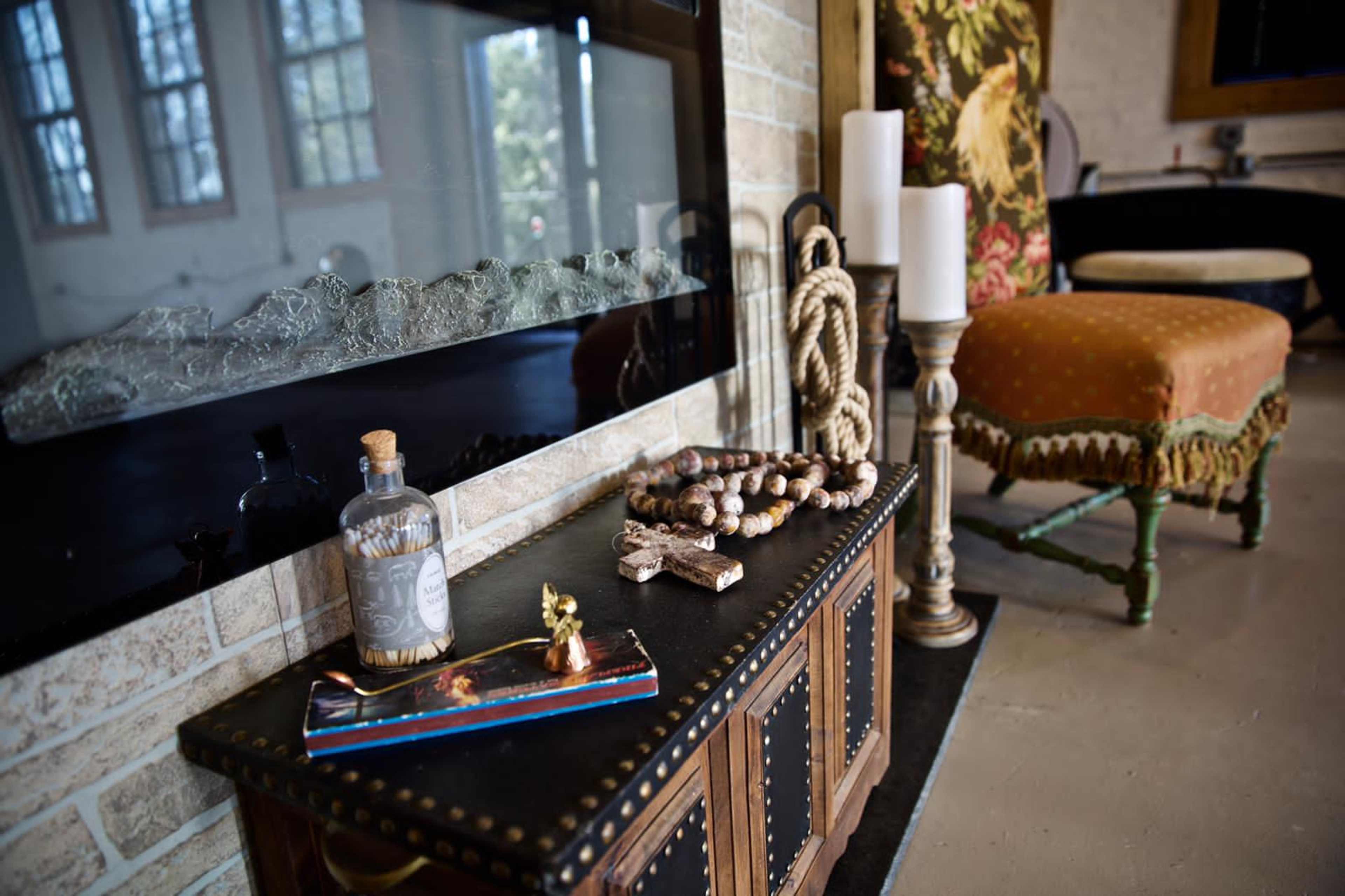 The scene features a decorative console table with a patterned runner, a glass bottle, a beaded necklace, candles, and an ornate stool beside a stone fireplace.