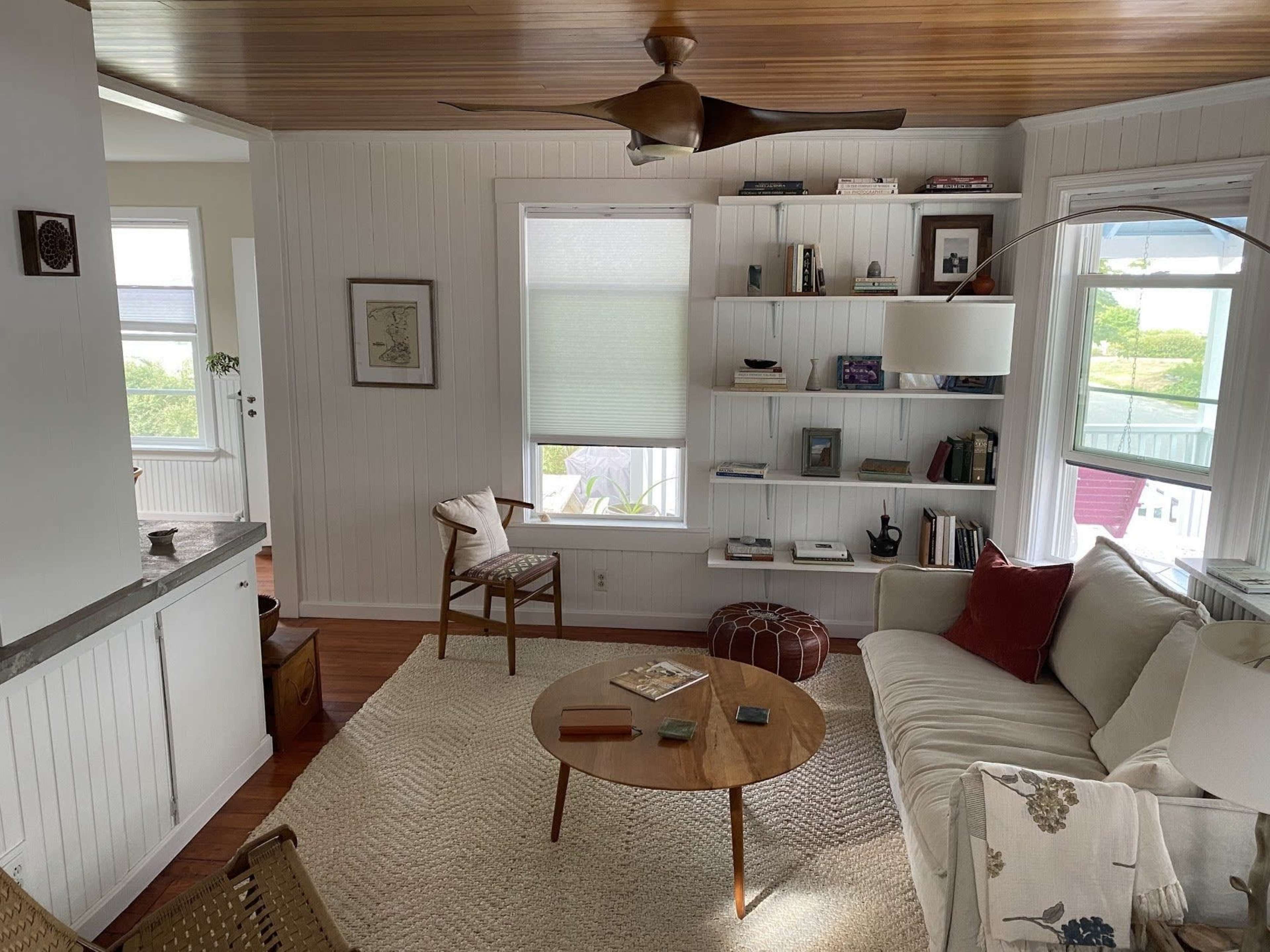 A cozy living room featuring a white sofa, a wooden coffee table, a bookshelf filled with books, and large windows allowing natural light to enter.
