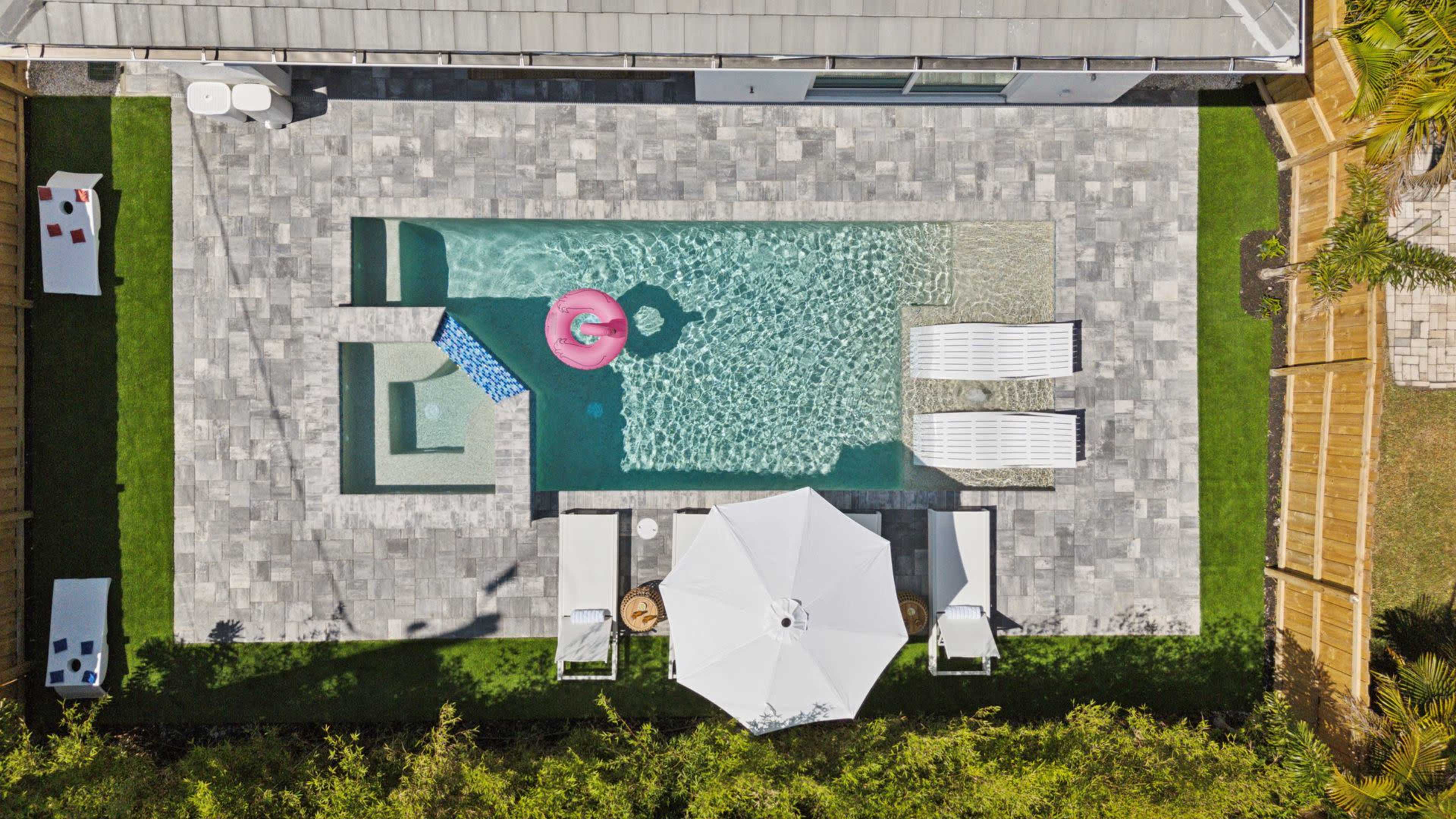 An aerial view shows a rectangular pool surrounded by stone tiles, with a pink float in the water, lounge chairs, an umbrella, and a seating area nearby.
