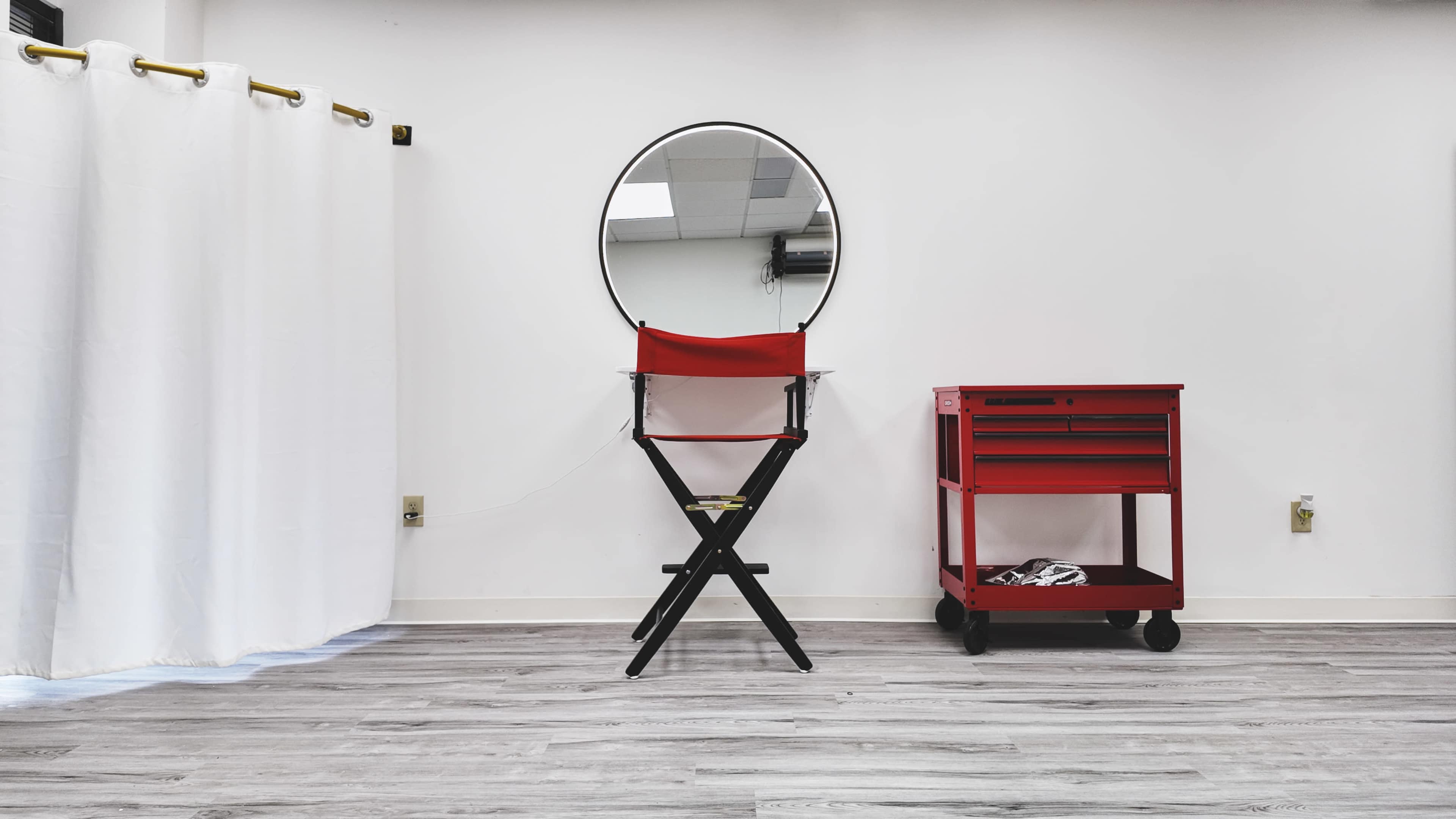 A circular mirror hangs above a foldable chair next to a red rolling cart in a minimalist room with white walls and wooden flooring.