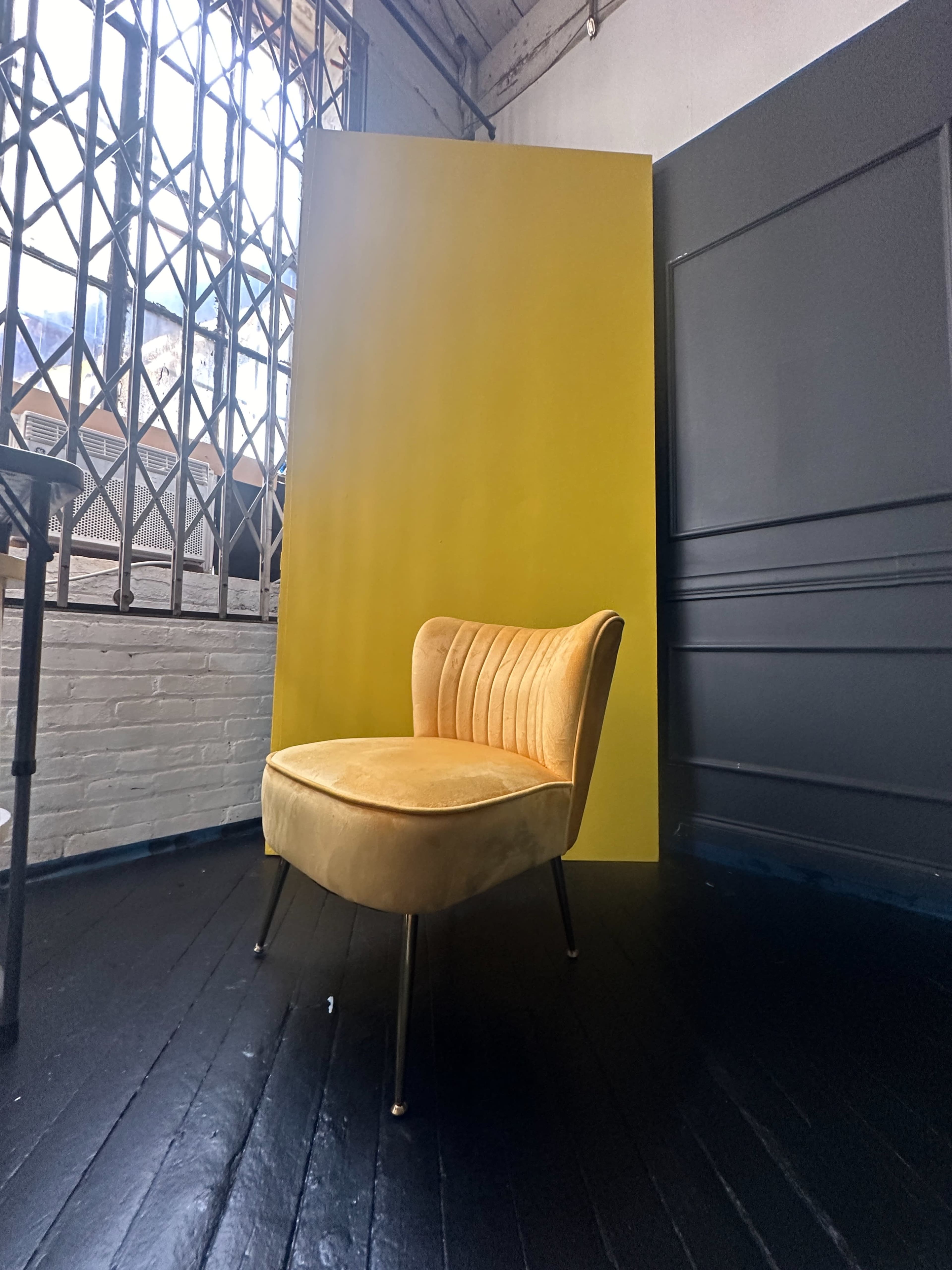 A yellow backdrop is positioned behind a tufted, mustard-colored chair in a bright, industrial space with a dark floor and large windows.