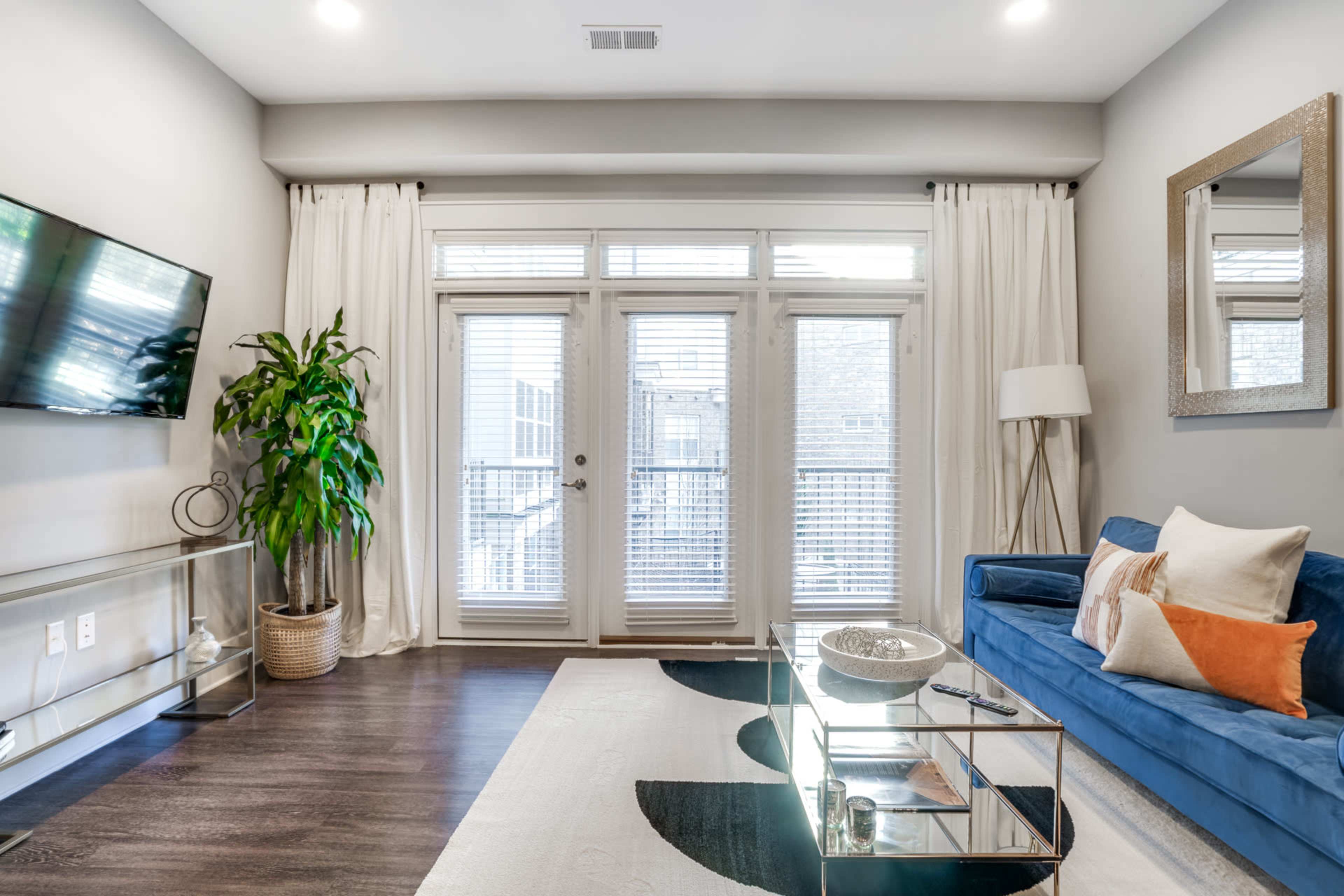 The living room features a blue sofa, a glass coffee table, a TV mounted on the wall, and large windows with white curtains.