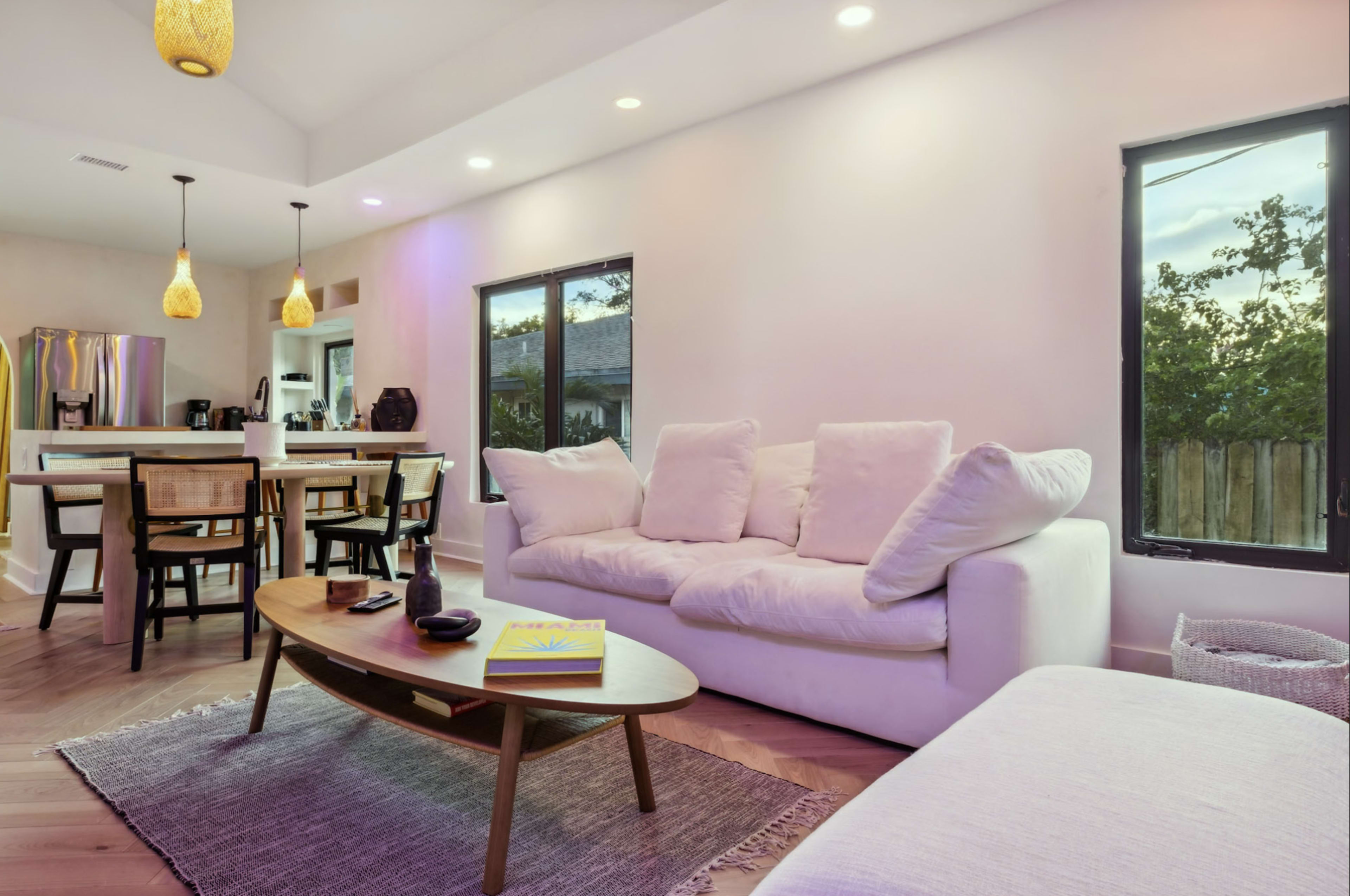 A modern living room features a white sofa, a wooden coffee table with a book, and large windows letting in natural light.