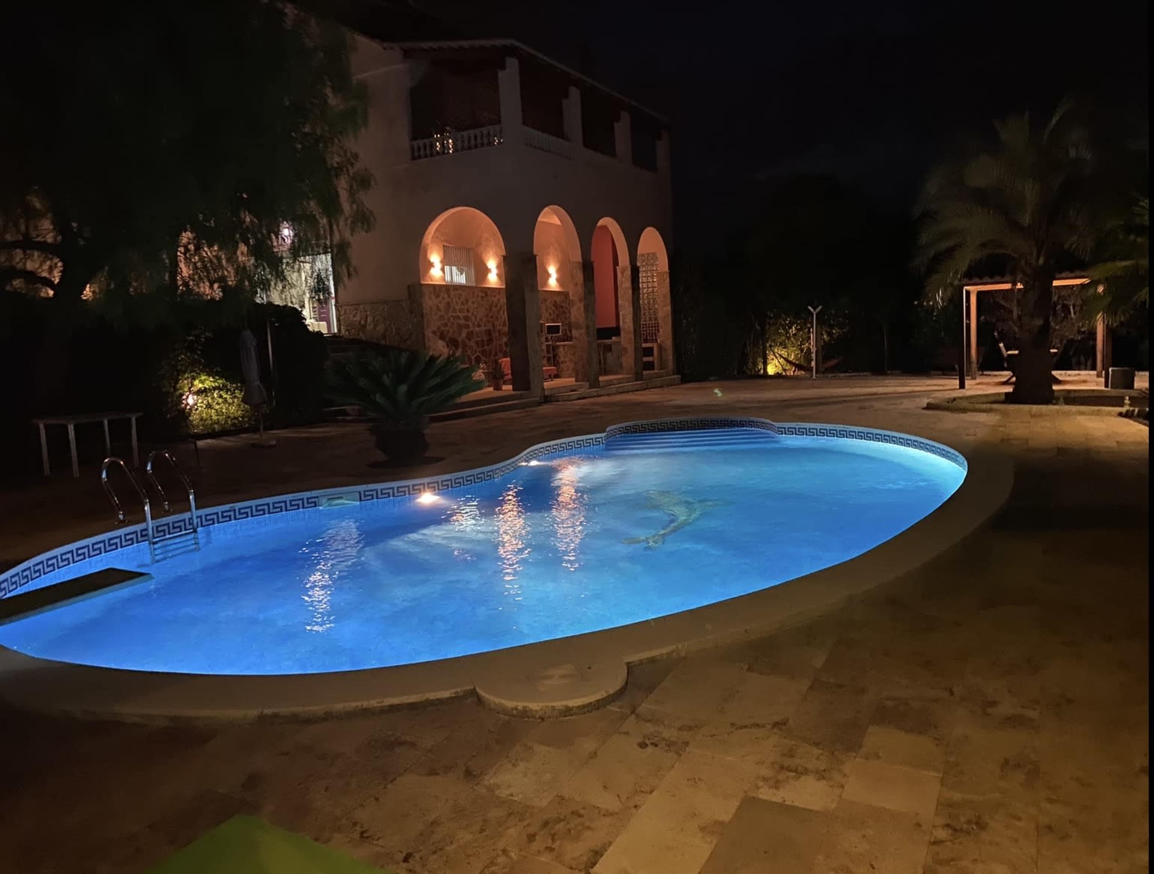 A lit swimming pool with a blue glow is situated in a well-manicured outdoor area featuring stone pathways and tropical plants, adjacent to a two-story building with arched windows.