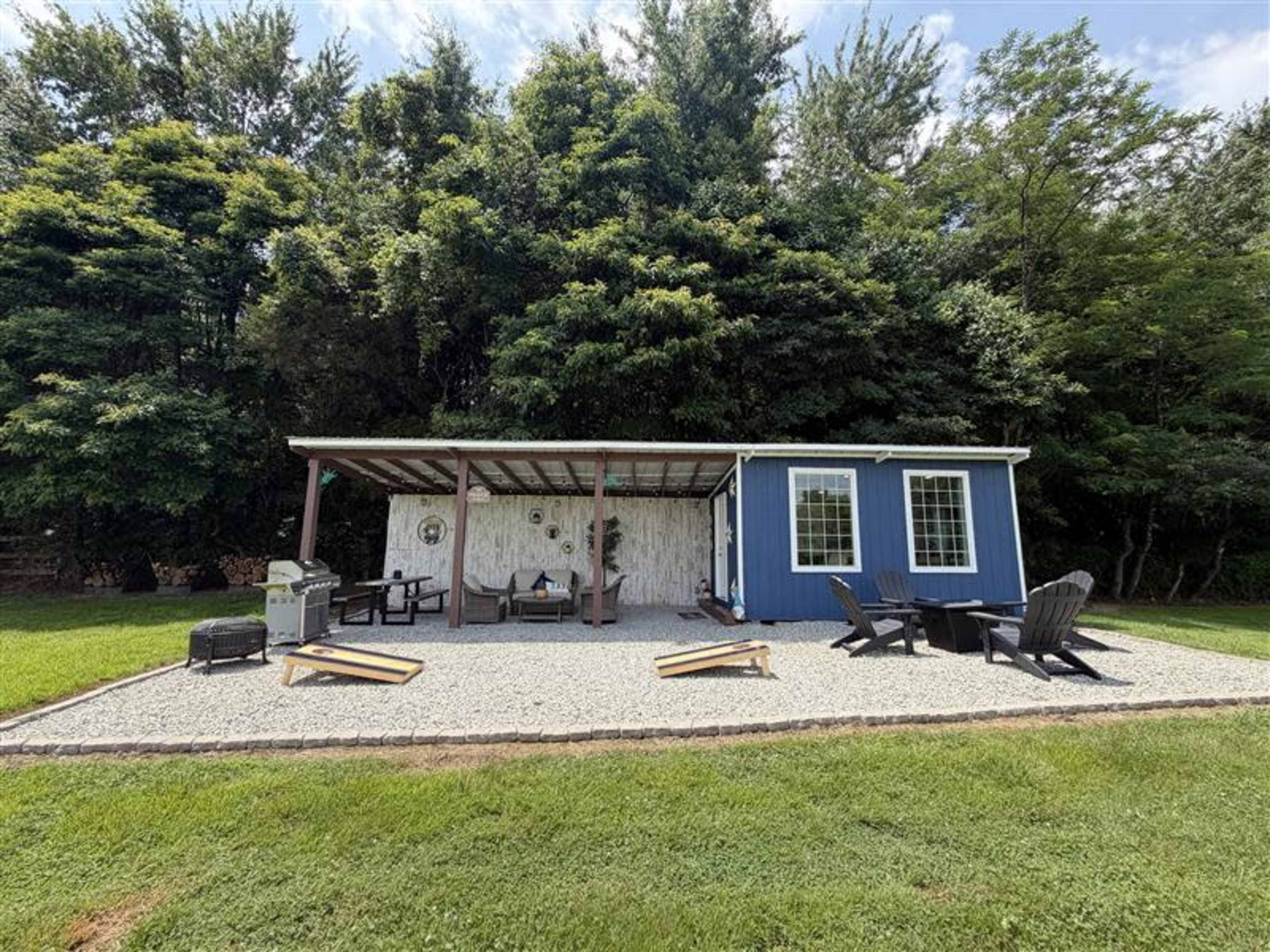 A covered outdoor seating area with a blue shed, wooden furniture, and a grill is situated on a gravel patio surrounded by greenery.
