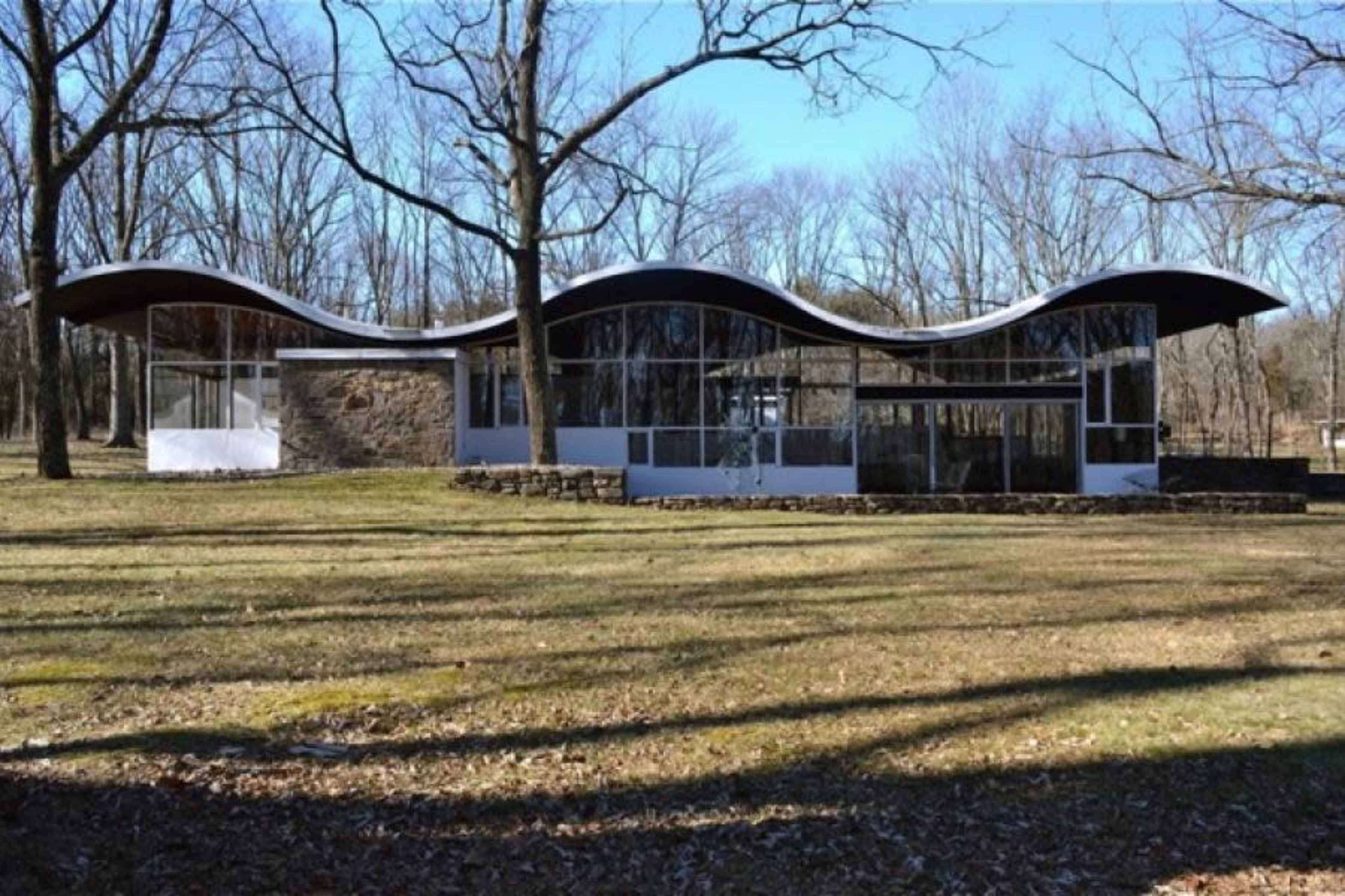 The image shows a modern, curvilinear building with large glass windows set among bare trees on a grassy landscape.