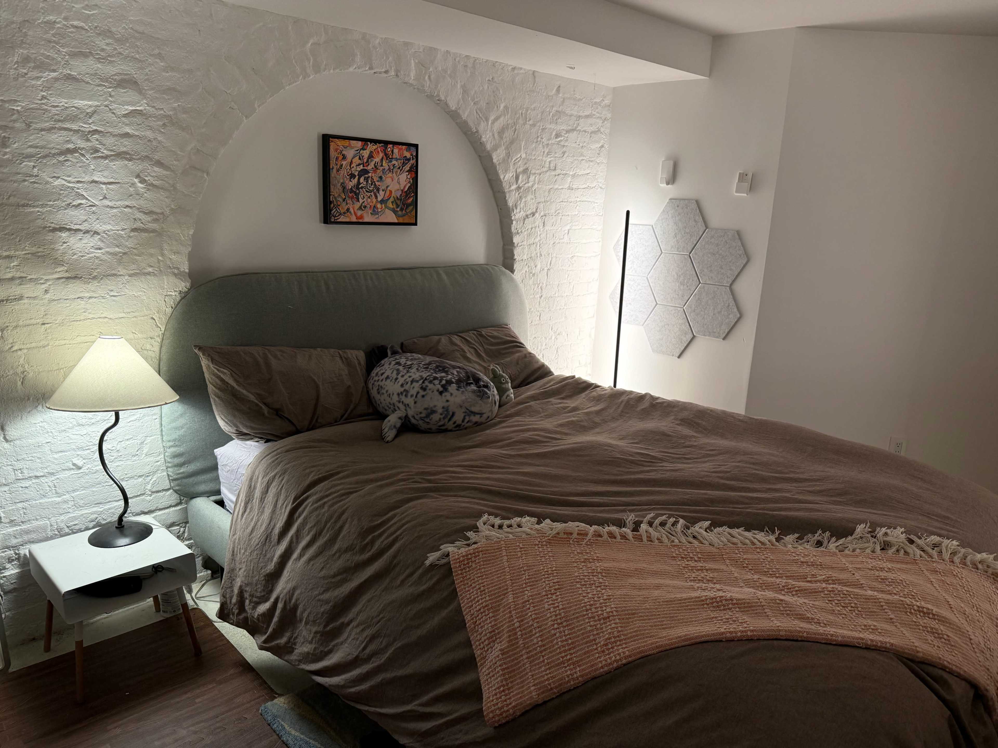 The image shows a cozy bedroom featuring a large bed with brown bedding, a plush pillow, and a bedside lamp beside a white brick wall.