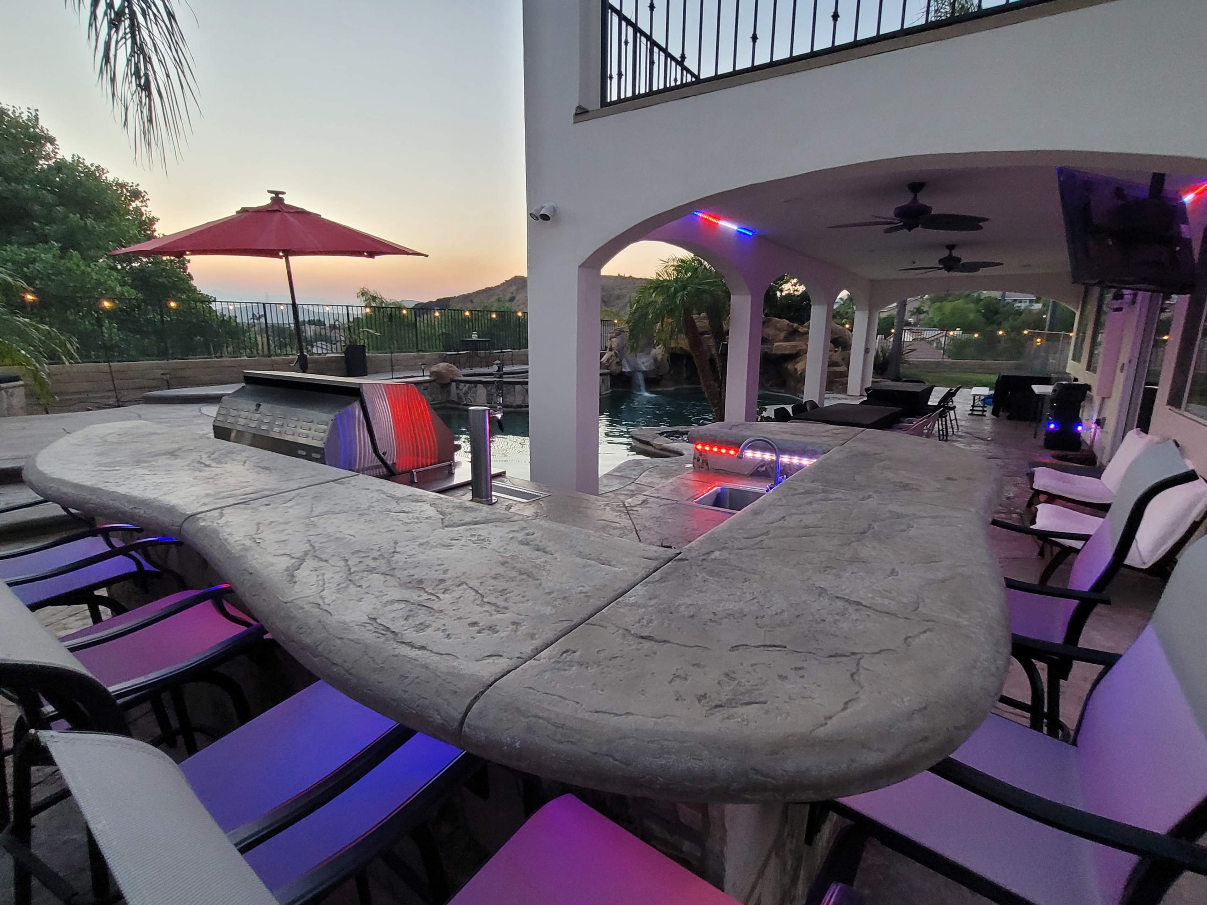 A curved outdoor bar area with seating, a grill, and a view of a pool surrounded by palm trees at sunset.