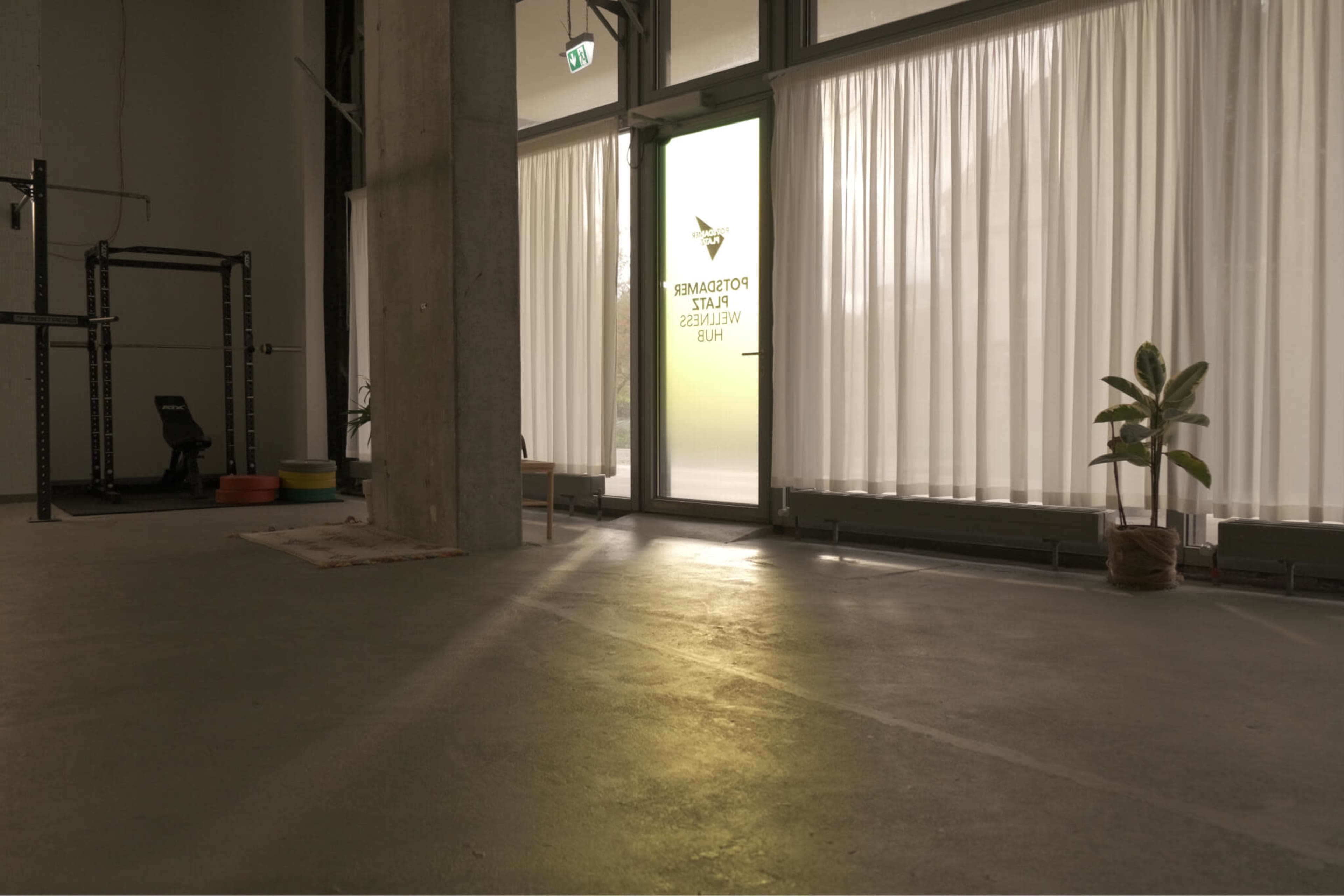 The image shows an empty interior space with large windows featuring sheer curtains and a door that has a logo, along with exercise equipment and a plant in the background.