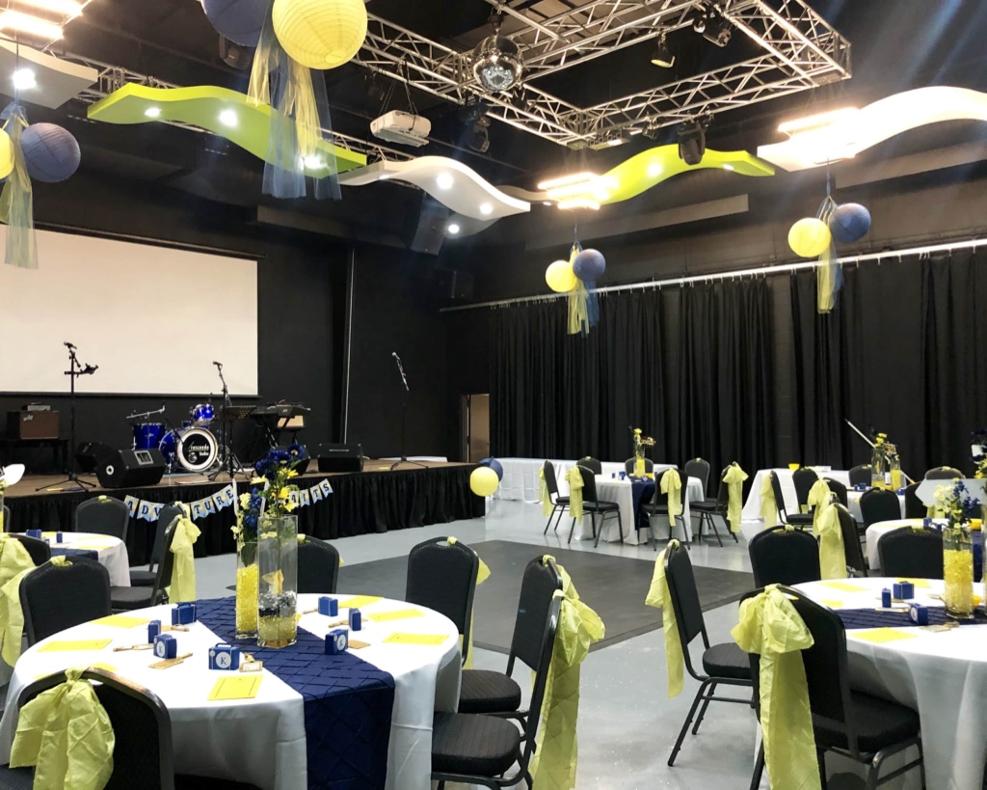 A decorated event space with tables set for a celebration, featuring balloons, banners, and a stage with musical instruments in the background.