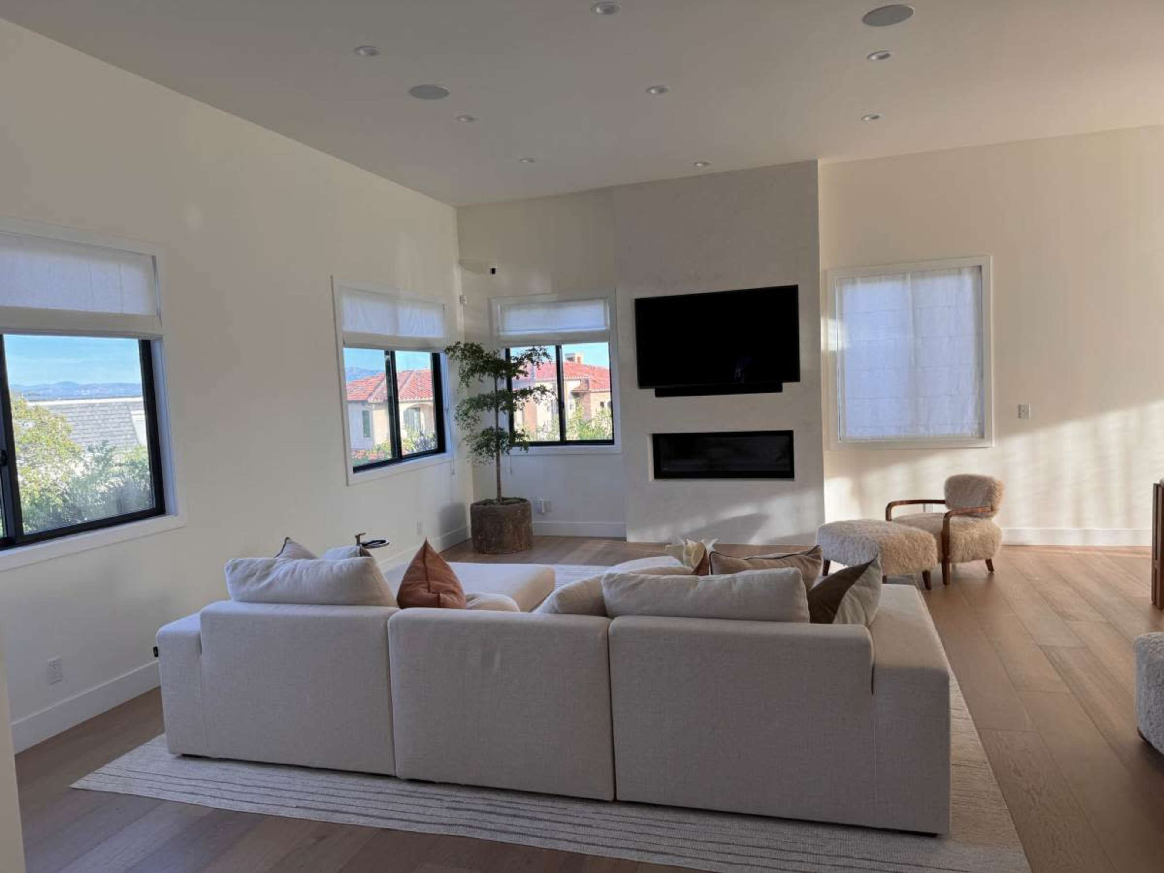 The image shows a bright living room featuring a large white sectional sofa, a mounted television, and large windows that provide a view of the outdoors.