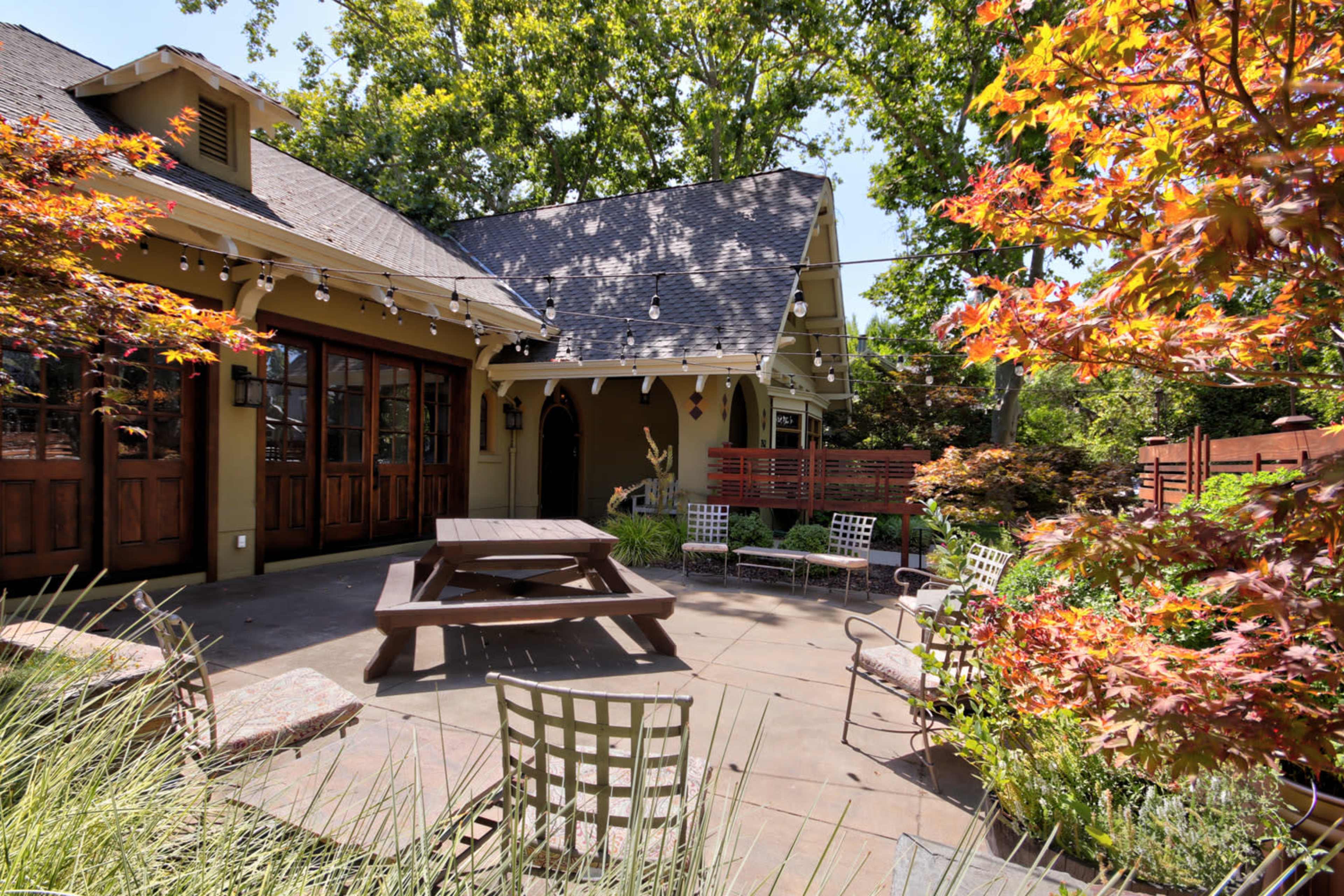 A patio area features a large picnic table surrounded by chairs and bordered by colorful landscaping and greenery.