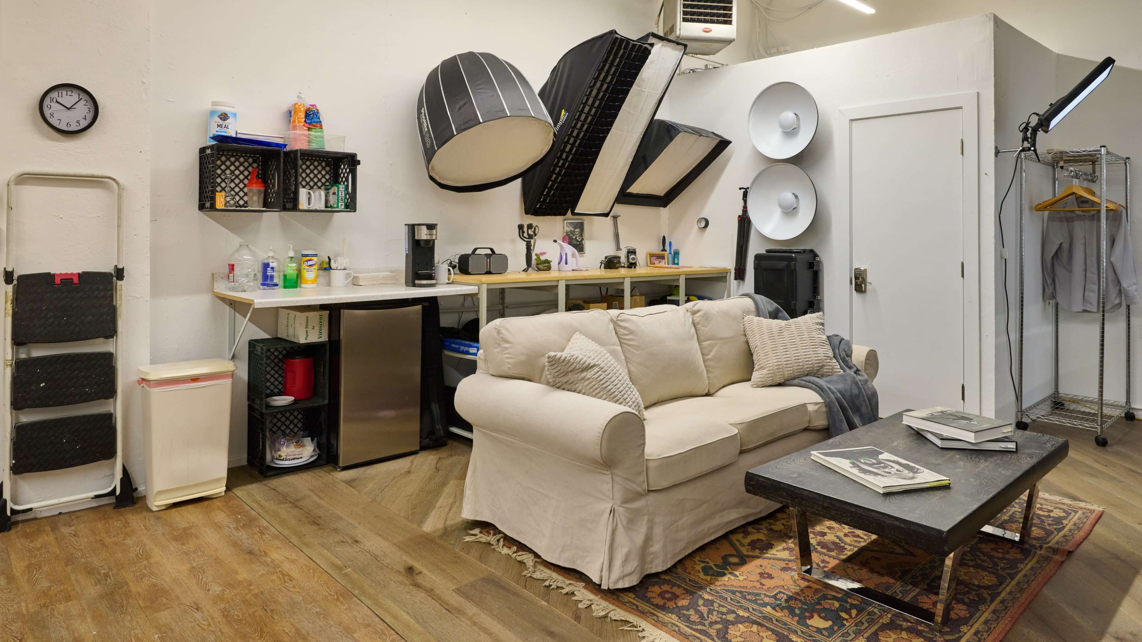A studio space with a sofa, coffee table, kitchen area, and various lighting equipment mounted on the walls.