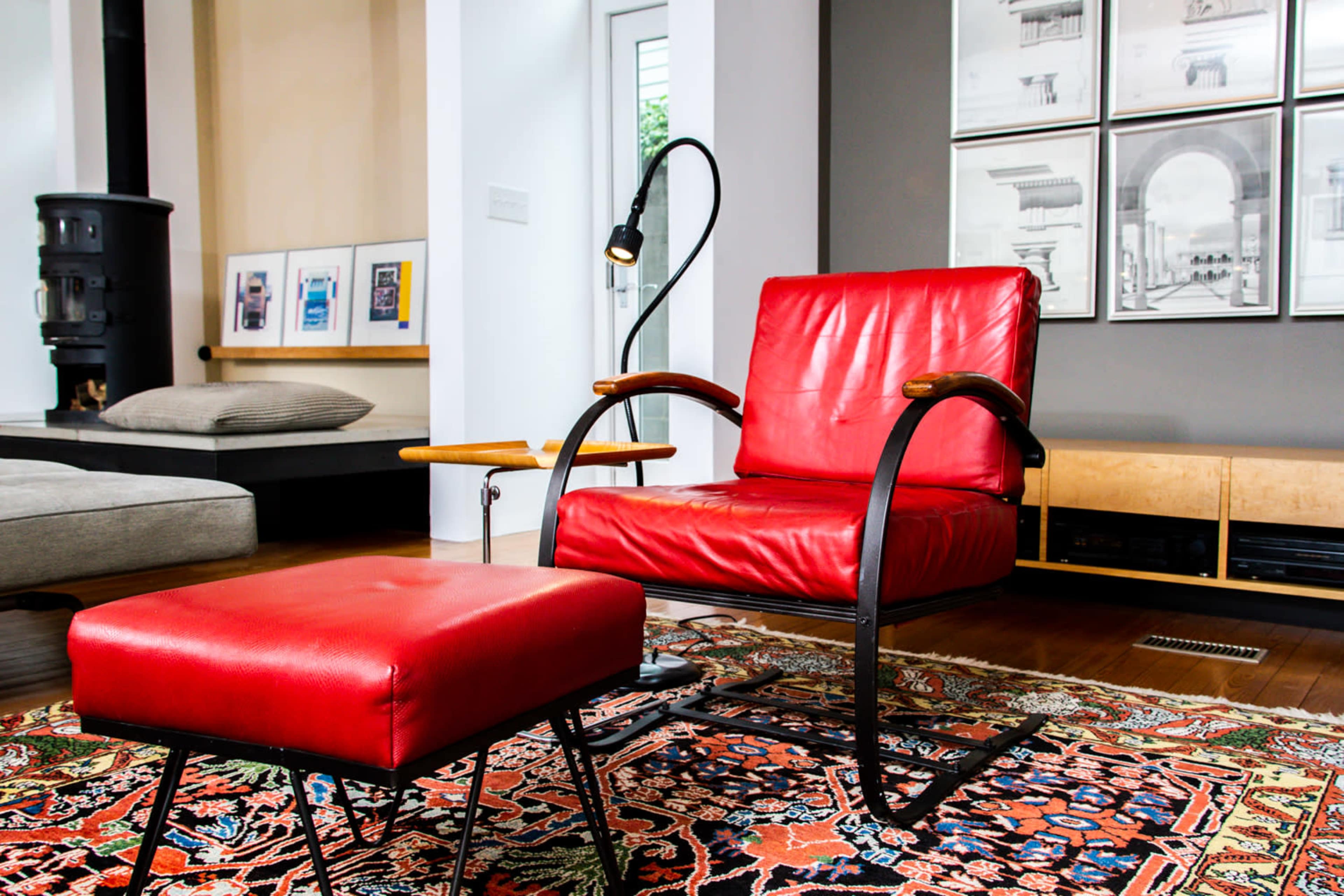 The image features a red leather armchair and ottoman on a patterned rug in a modern living room with framed artwork on the walls.