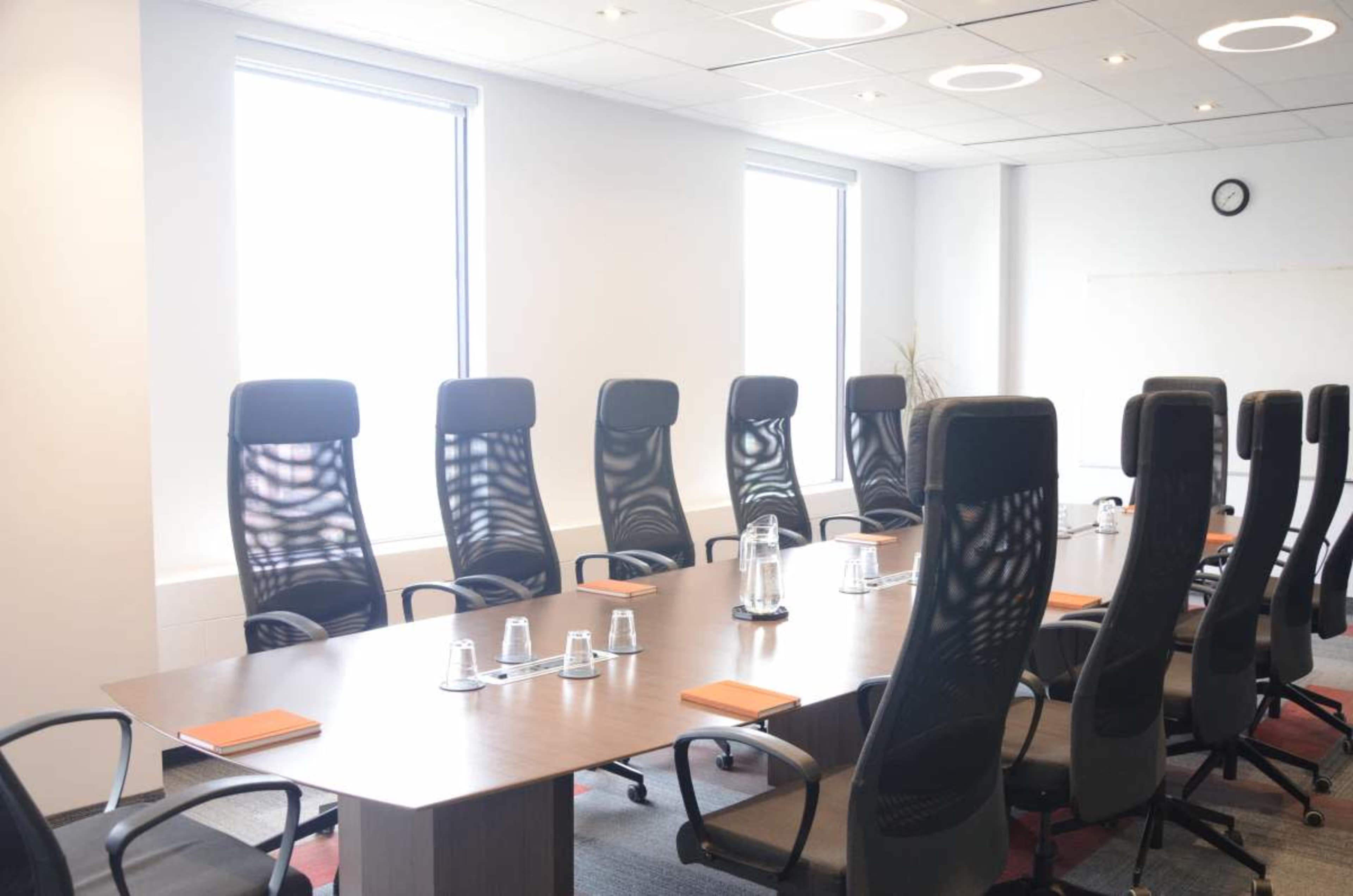 A large conference room features a long wooden table surrounded by ergonomic black chairs, with natural light streaming in from windows on one side.