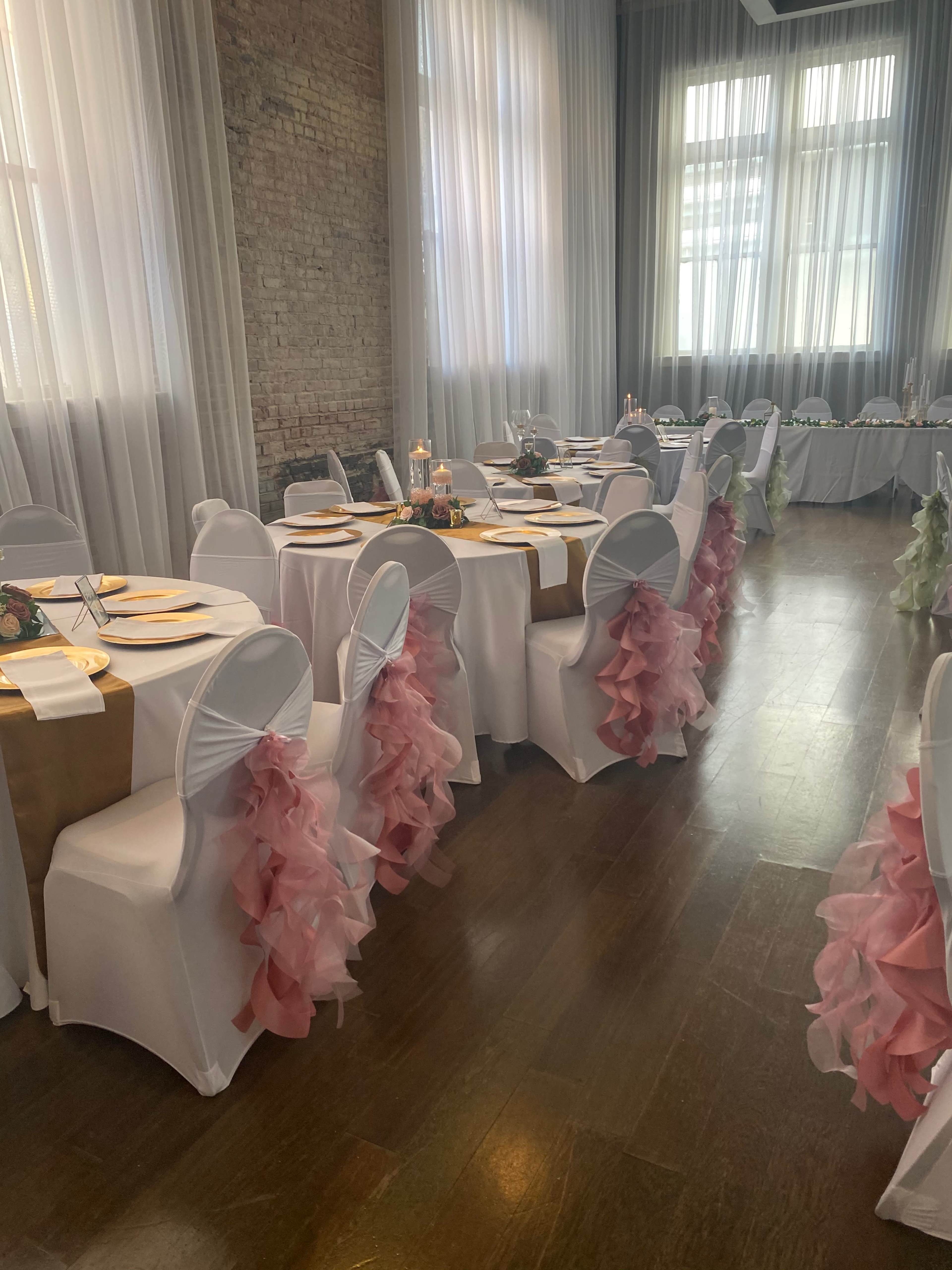 A decorated event space with tables covered in white tablecloths and pink and gold accents, featuring chairs draped with ruffled sashes, and large windows letting in natural light.