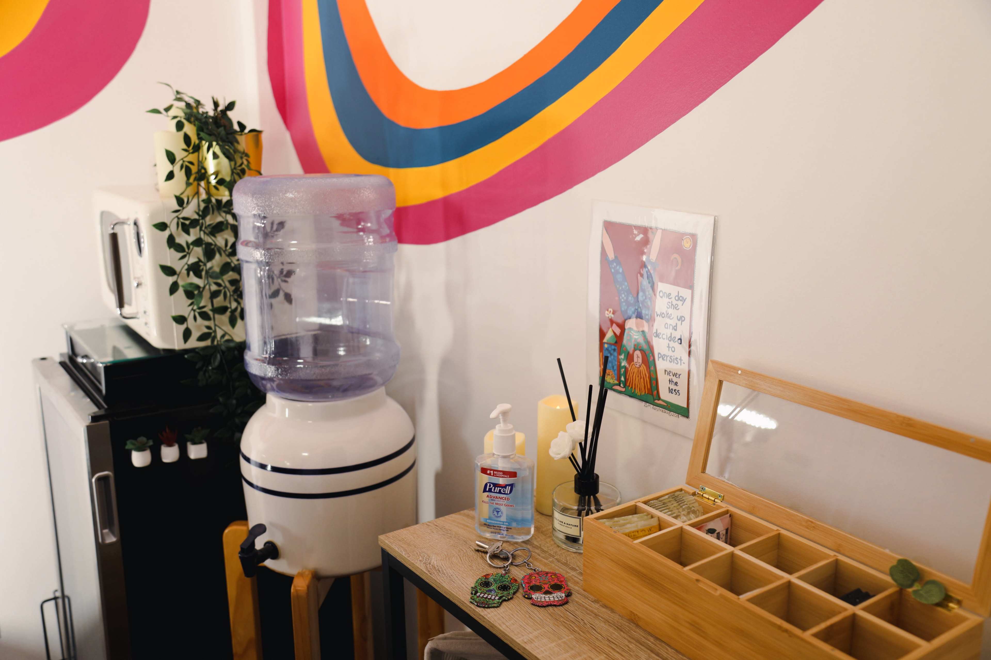 A water cooler stands beside a wooden table displaying a box of items, hand sanitizer, and decorative accessories against a wall with colorful stripes.