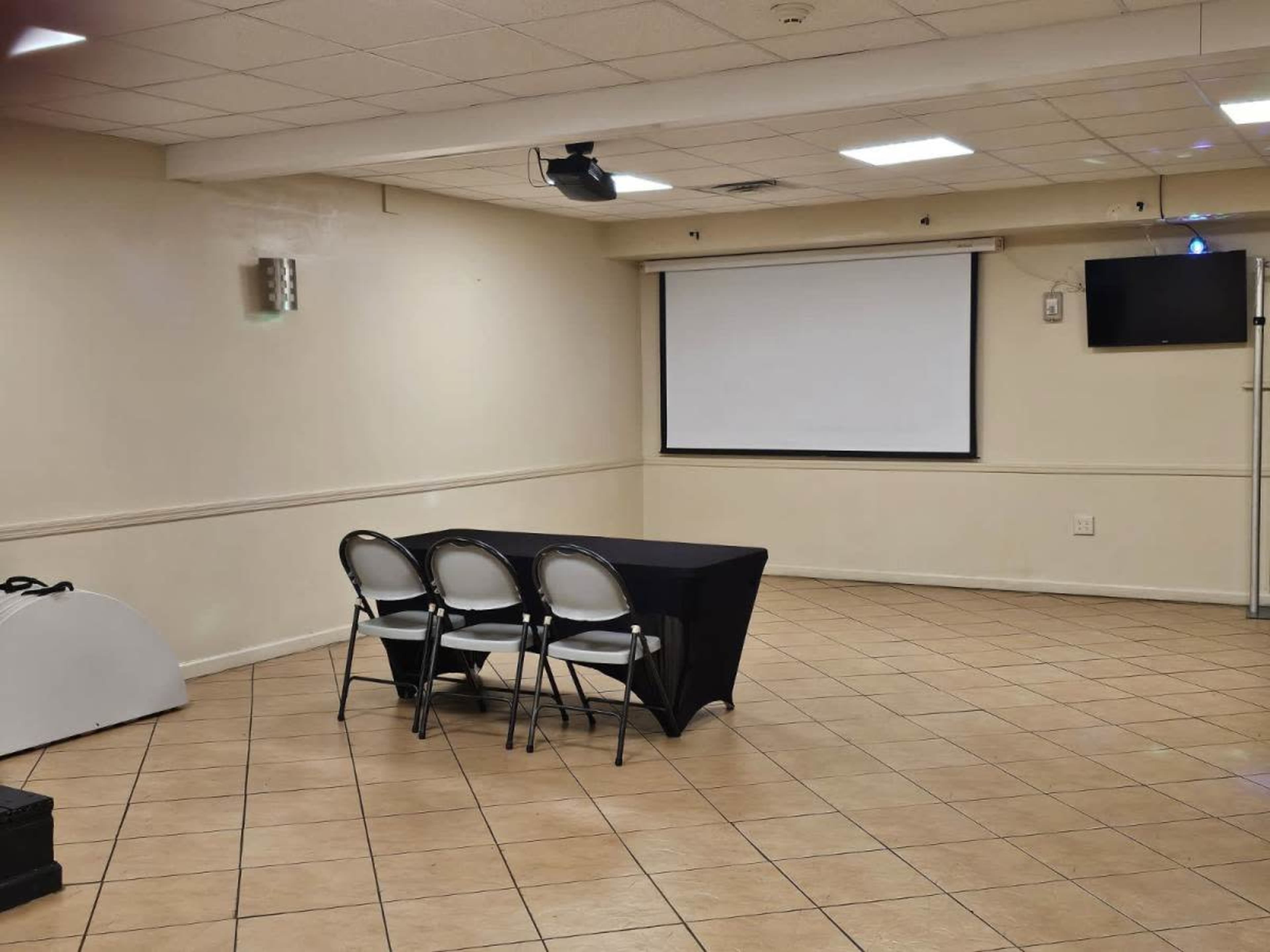 A sparsely furnished meeting room with a black table and three chairs facing a blank projector screen.