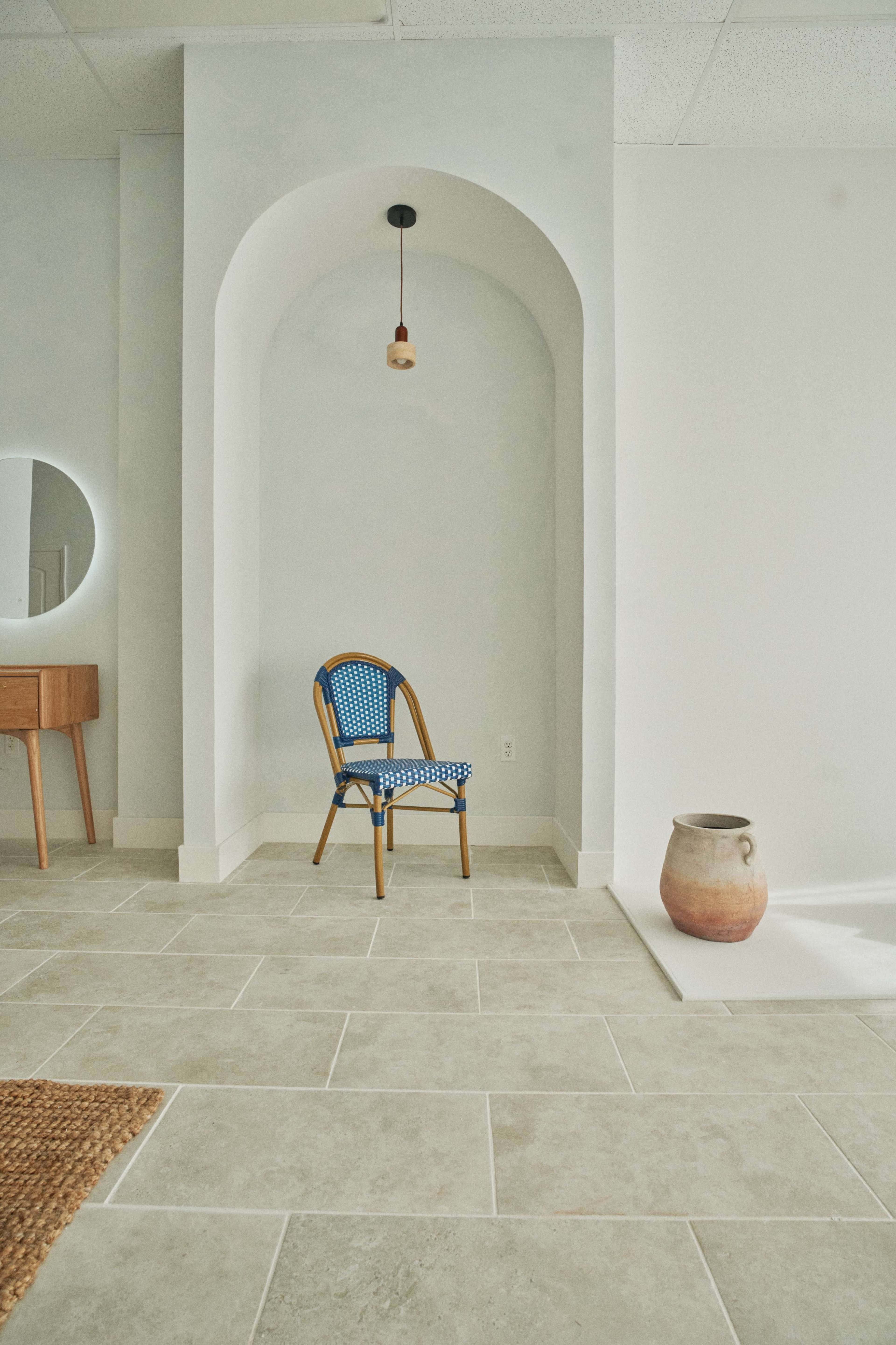 The image features a minimalist interior space with a blue woven chair placed against a light-colored wall that has an archway, alongside a decorative ceramic pot and a round mirror.