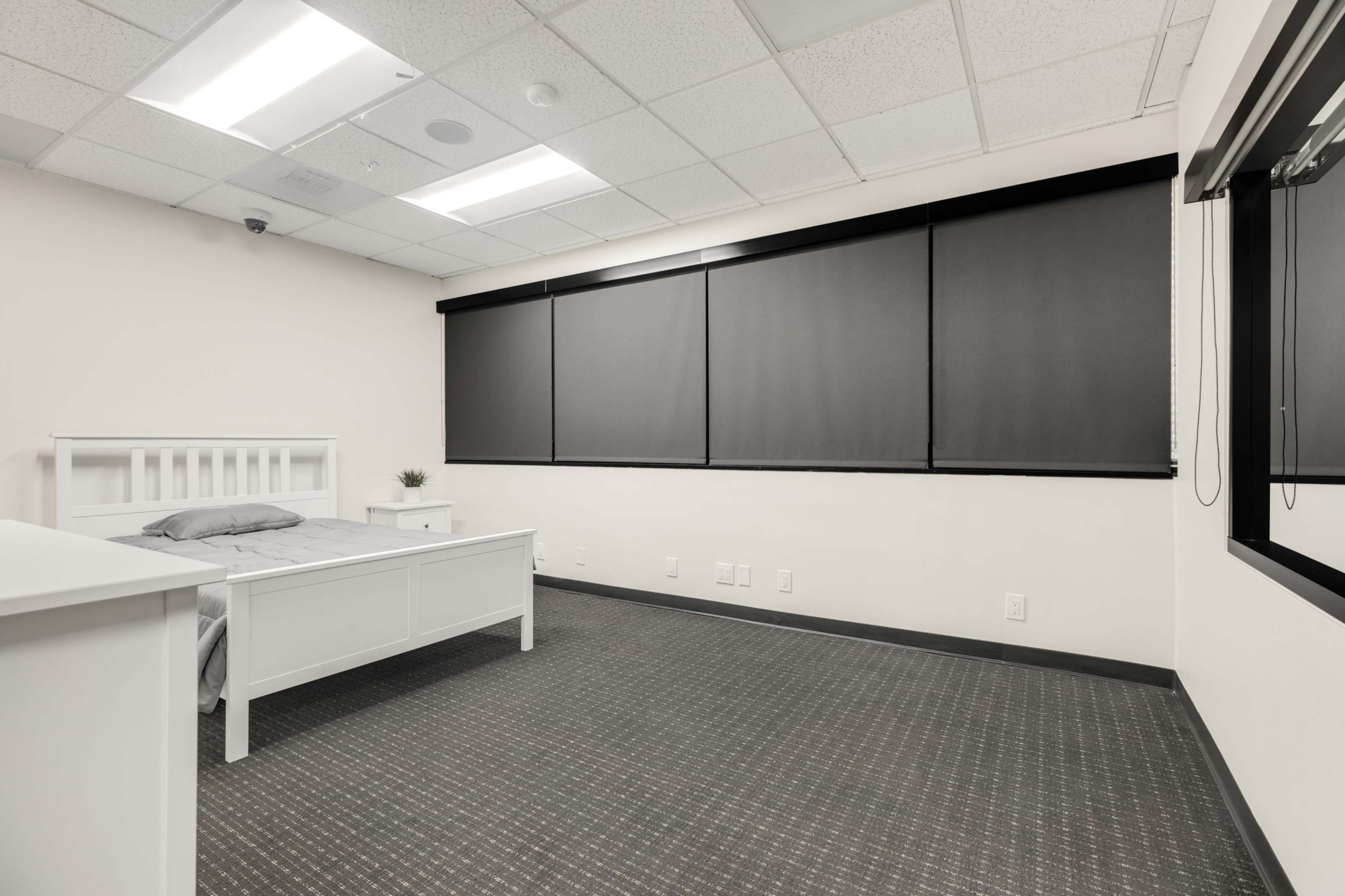 The image shows a minimalist bedroom with a white bed frame, gray bedding, and dark window shades.