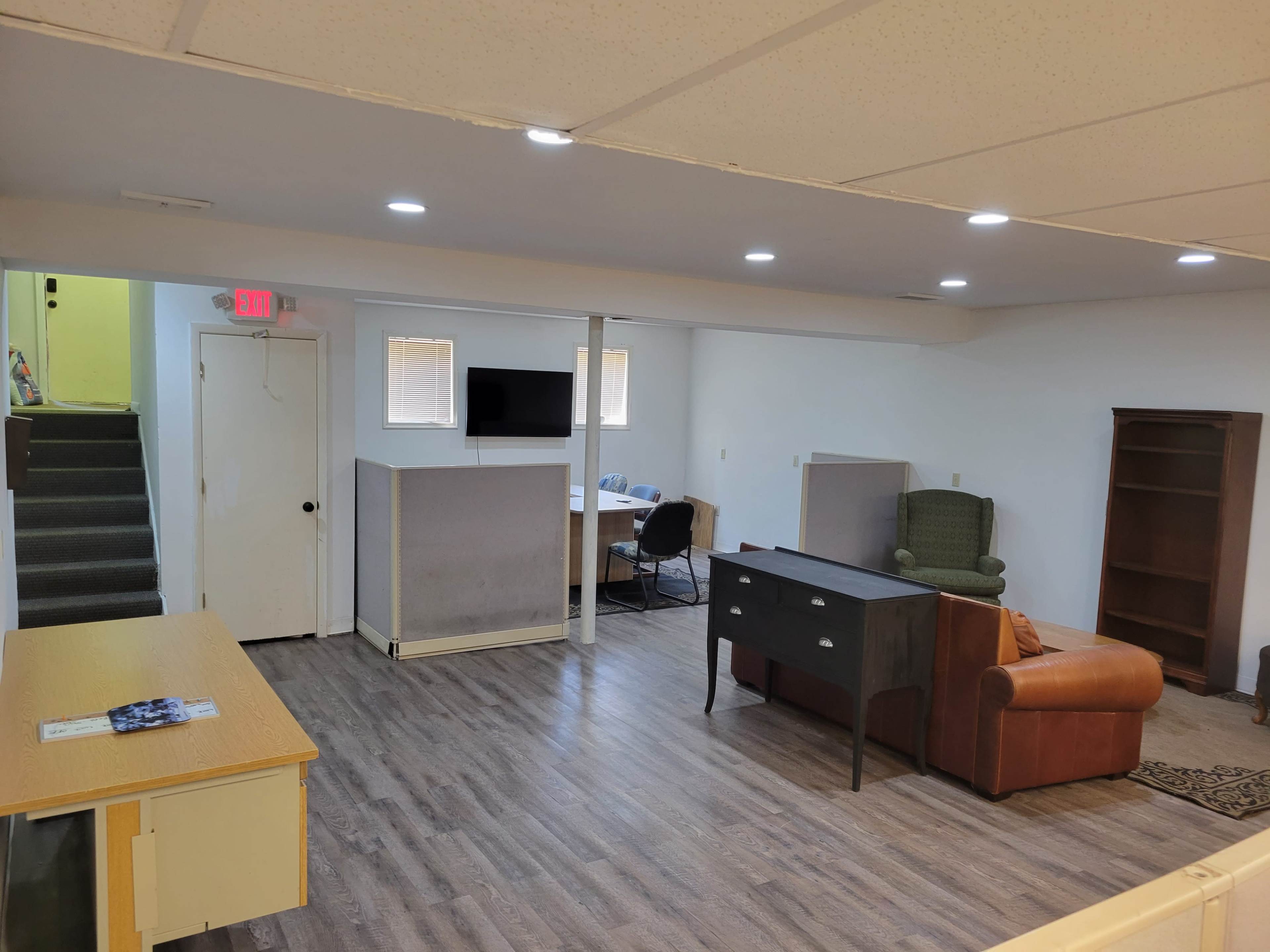 A spacious basement area with a desk, chair, sofa, and bookshelves, along with a staircase leading to another level.