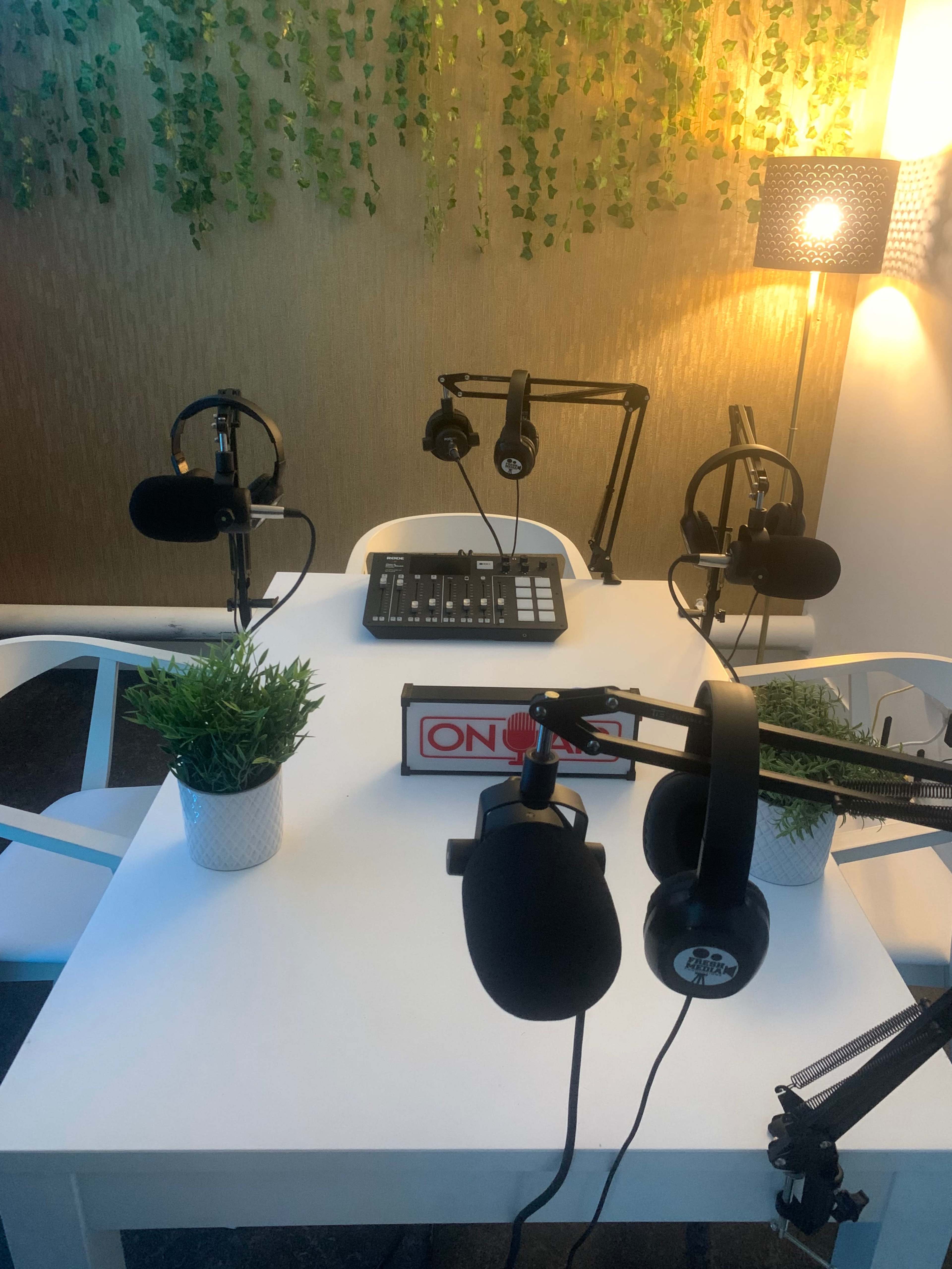 A podcasting studio setup with a white table, four microphones on adjustable arms, a mixing console, and decorative plants.