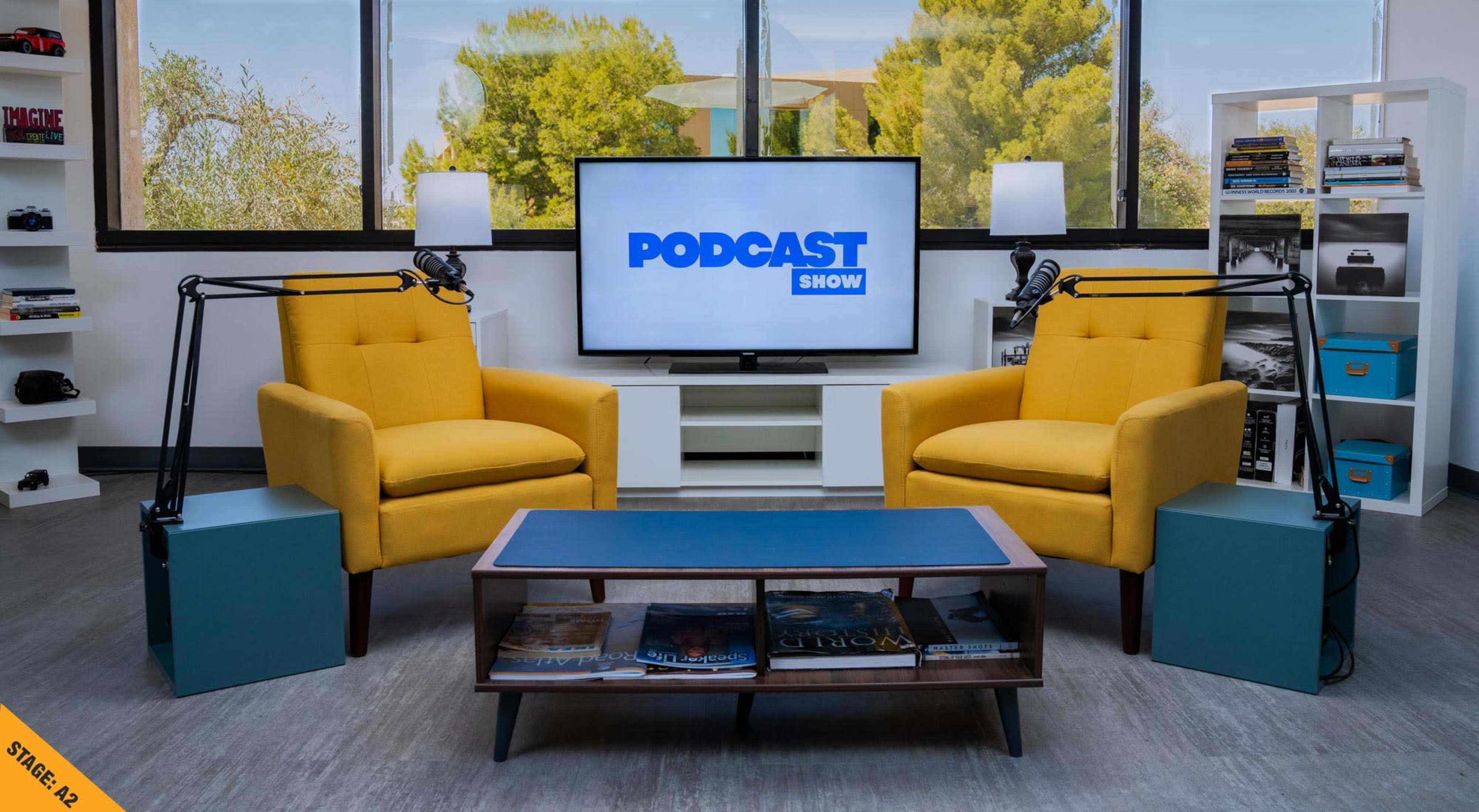 The image shows a modern podcasting setup featuring two yellow armchairs facing a coffee table, with a television displaying the text "PODCAST SHOW" and a backdrop of greenery outside.