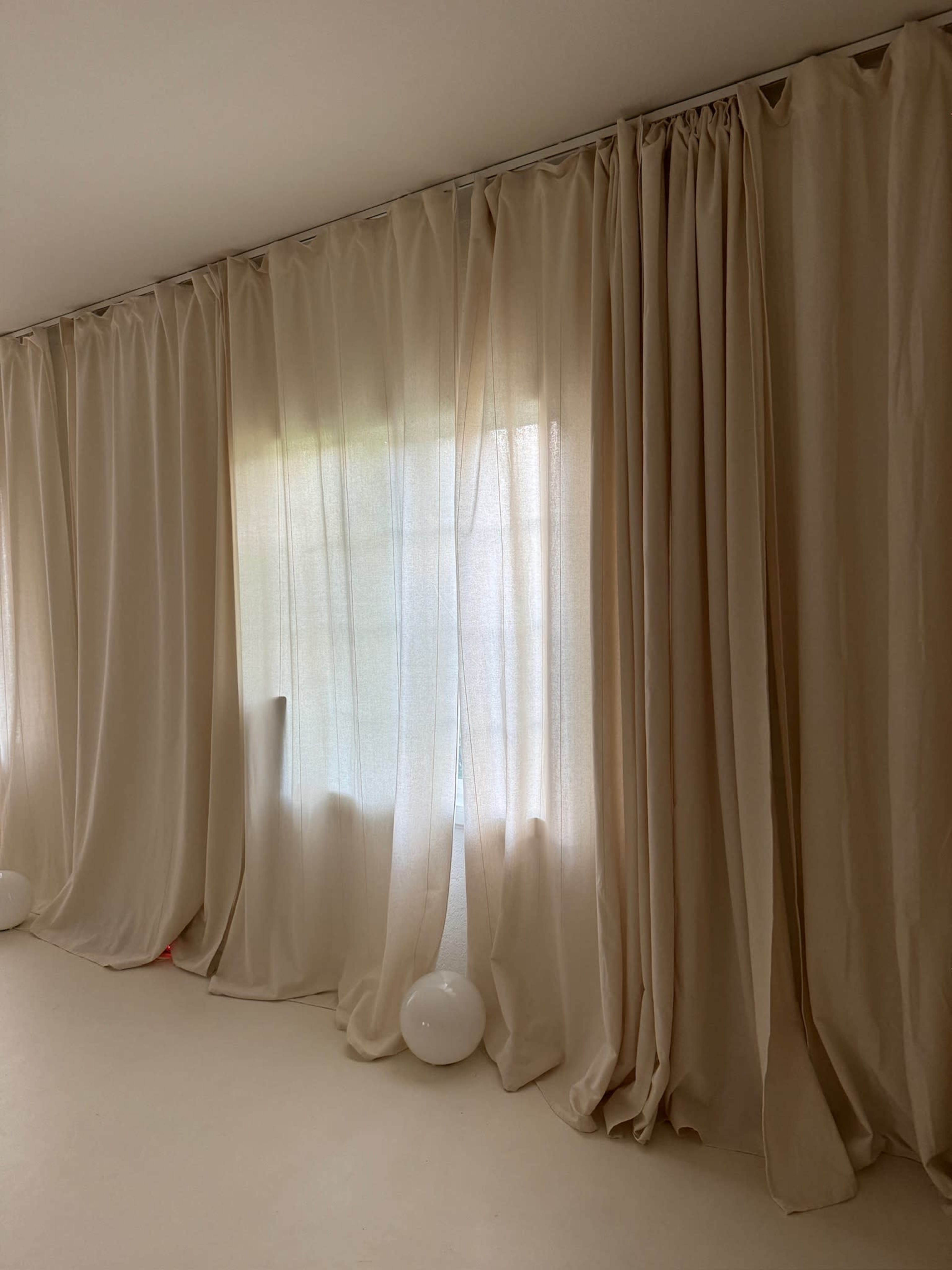 The image shows a wall with flowing beige curtains hanging over a window, partially diffusing natural light.