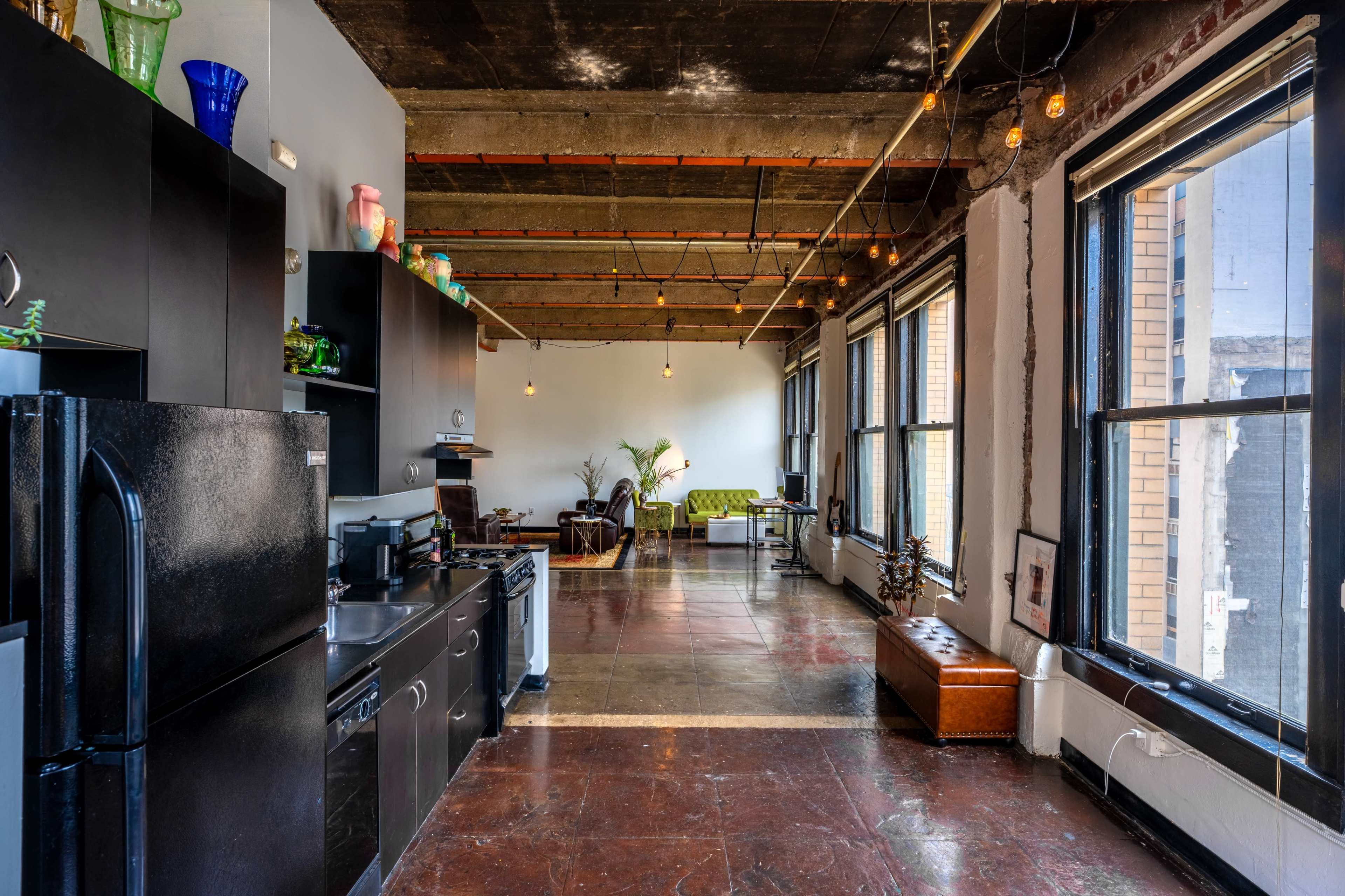 A modern industrial-style kitchen and living space with large windows, exposed ceiling beams, and dark cabinetry.