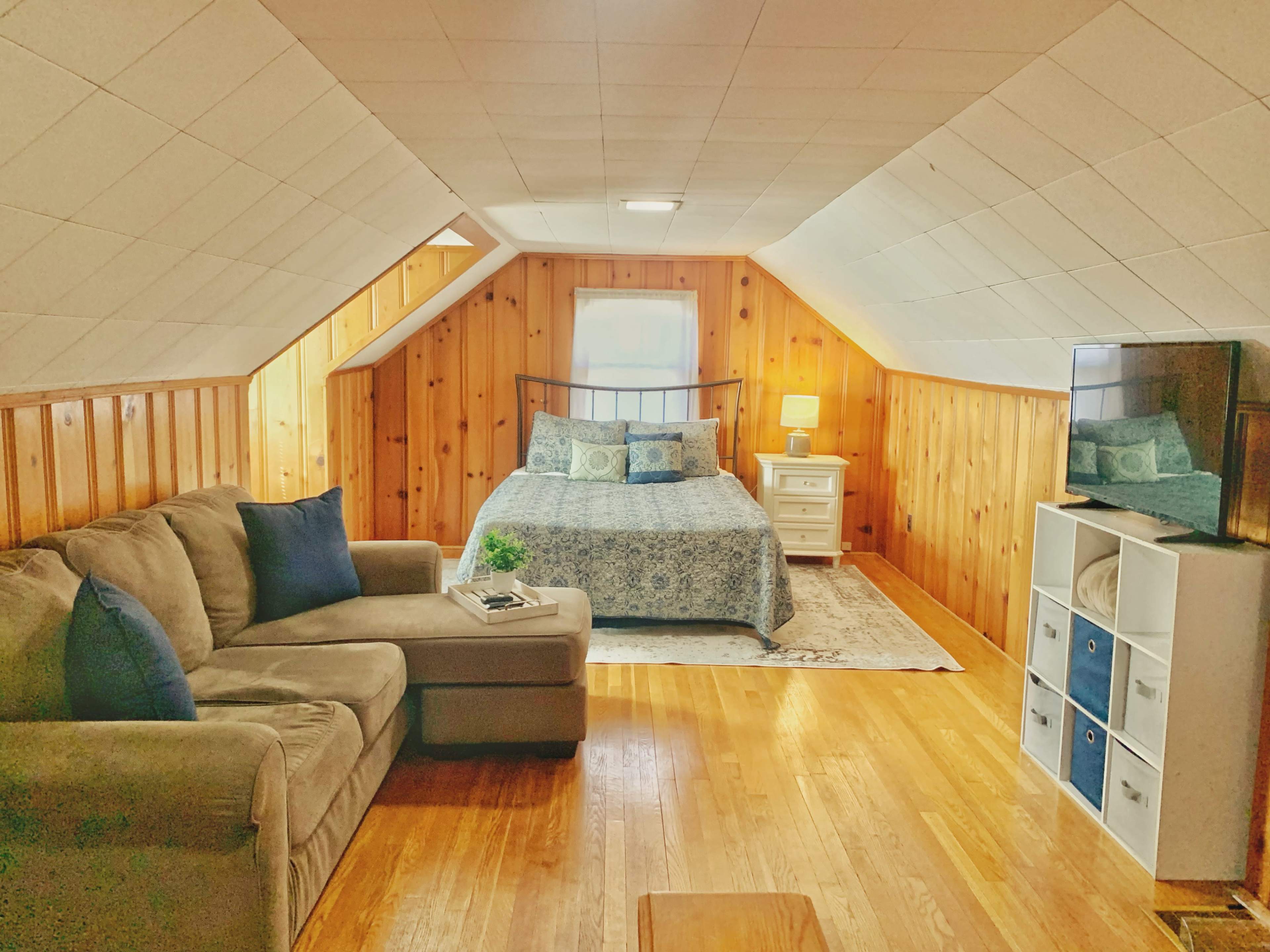 An inviting attic bedroom featuring a bed with a blue patterned quilt, a beige sofa, a side table with a lamp, and a TV on a stand.