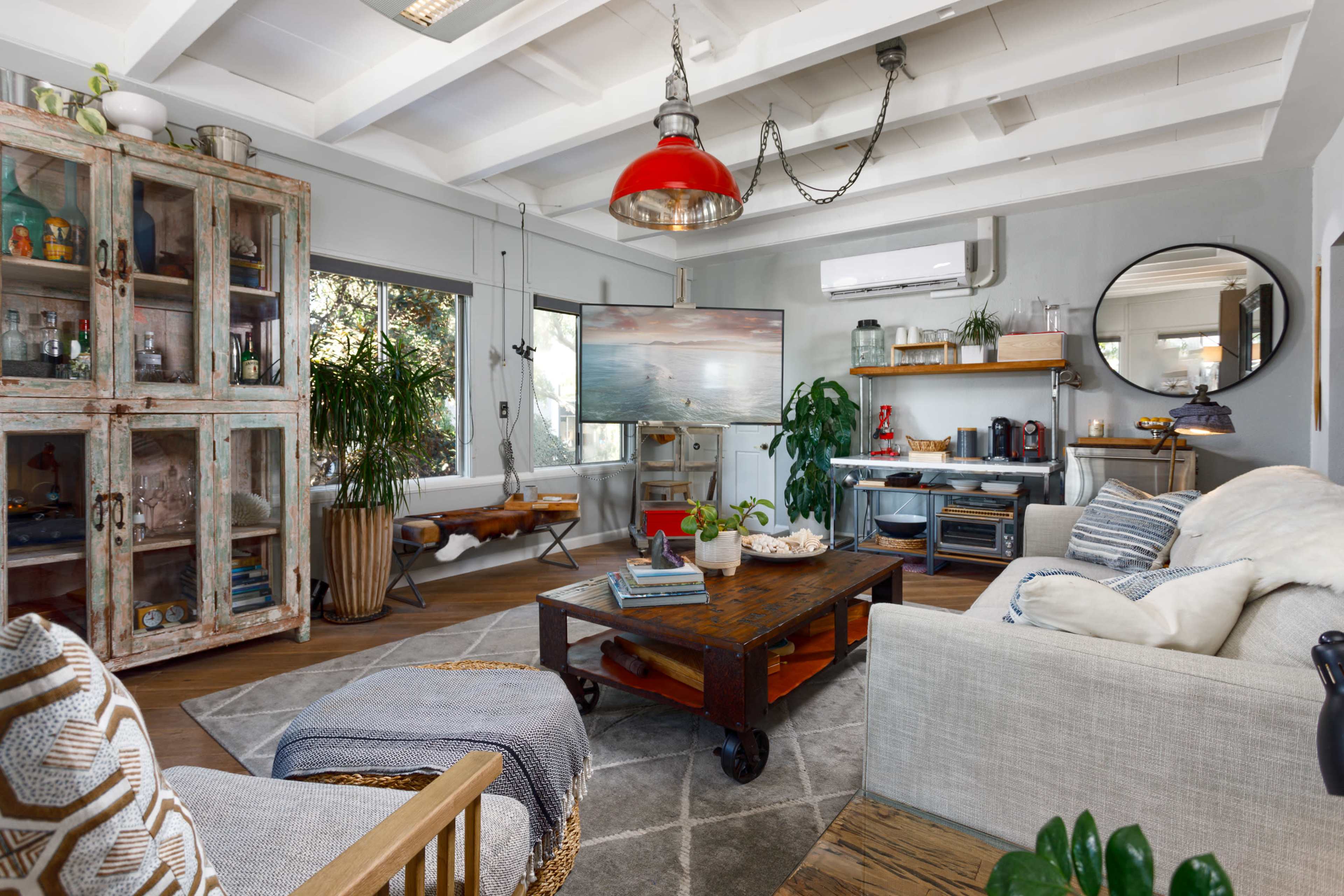A cozy living room featuring a large window, a rustic wooden coffee table, a glass-fronted cabinet, and various plants arranged throughout the space.