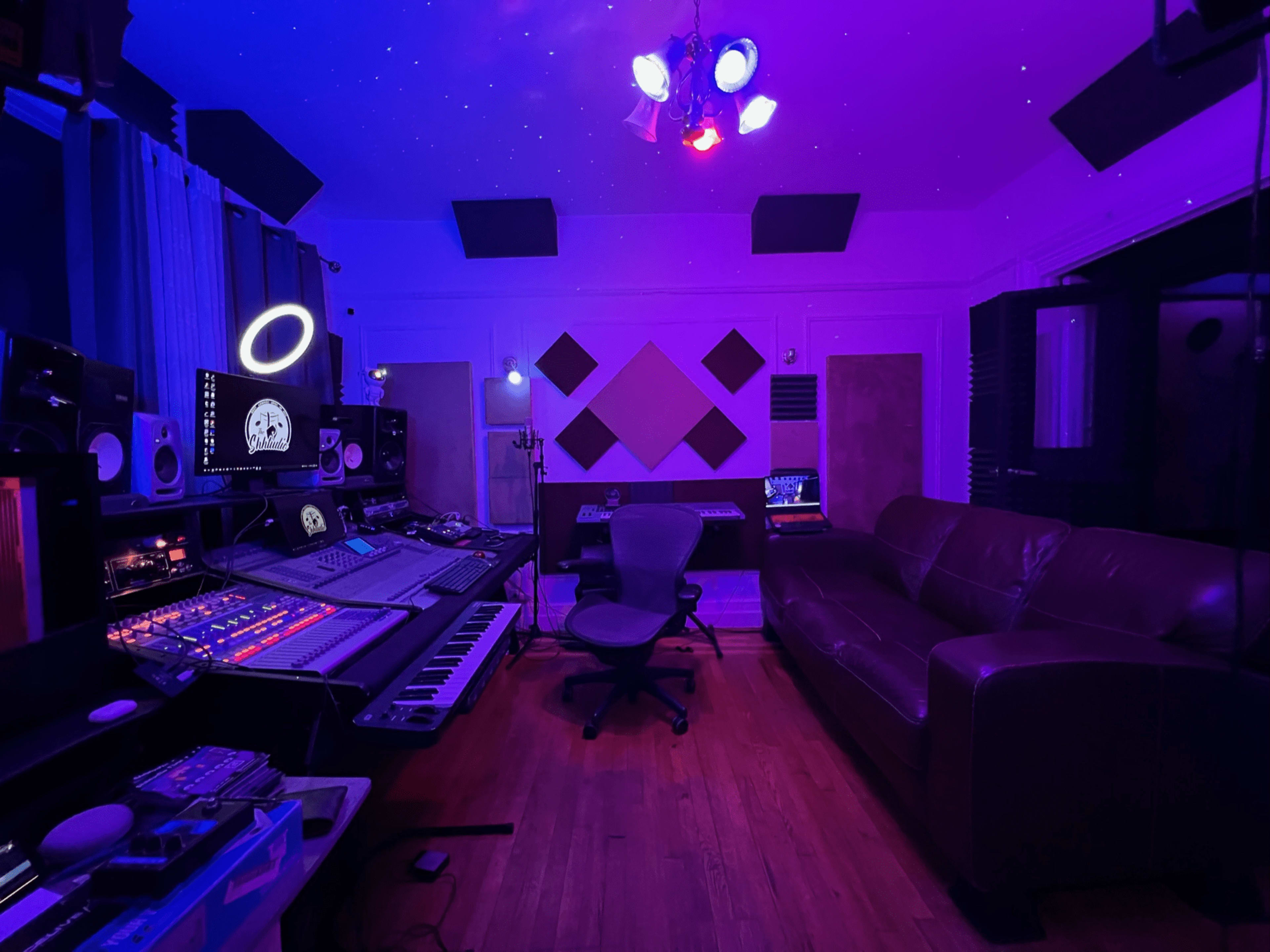 A music studio with a mixing console, computer equipment, and a sofa, all illuminated by purple and blue lighting.