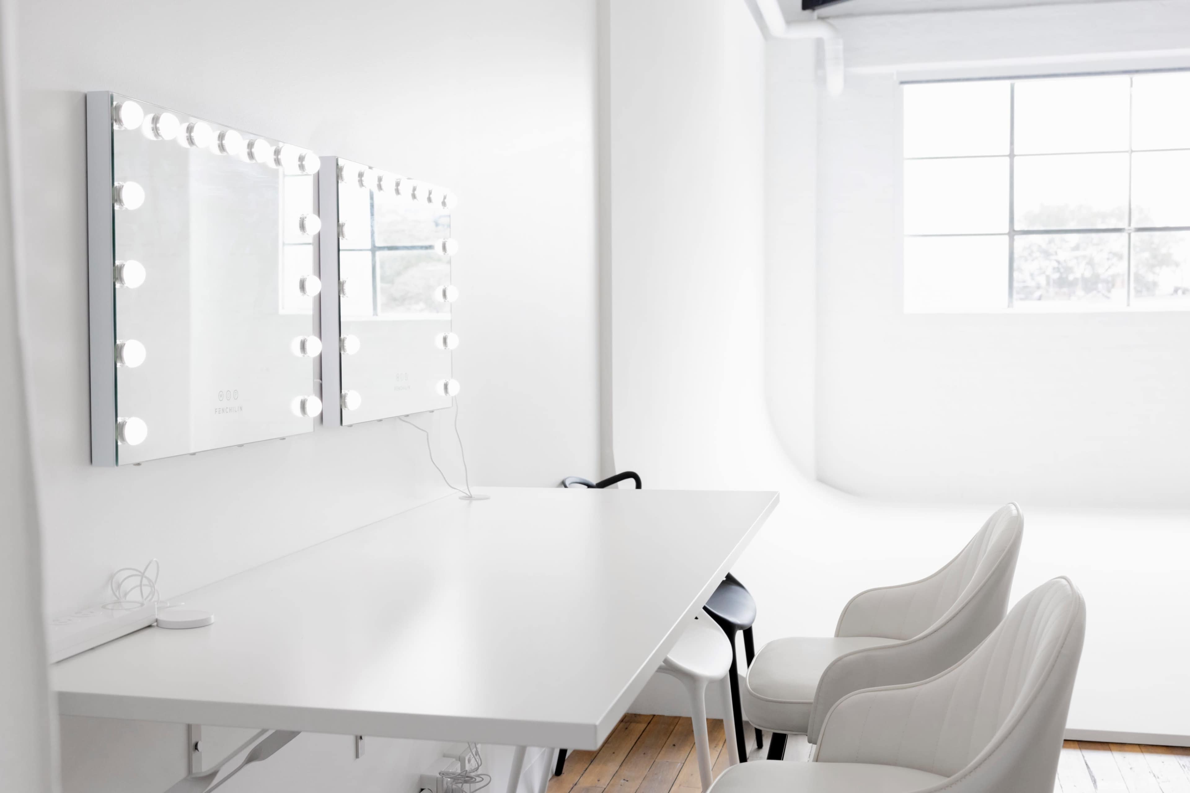 A bright, minimalistic dressing room with two lighted mirrors above a white table and two white chairs.