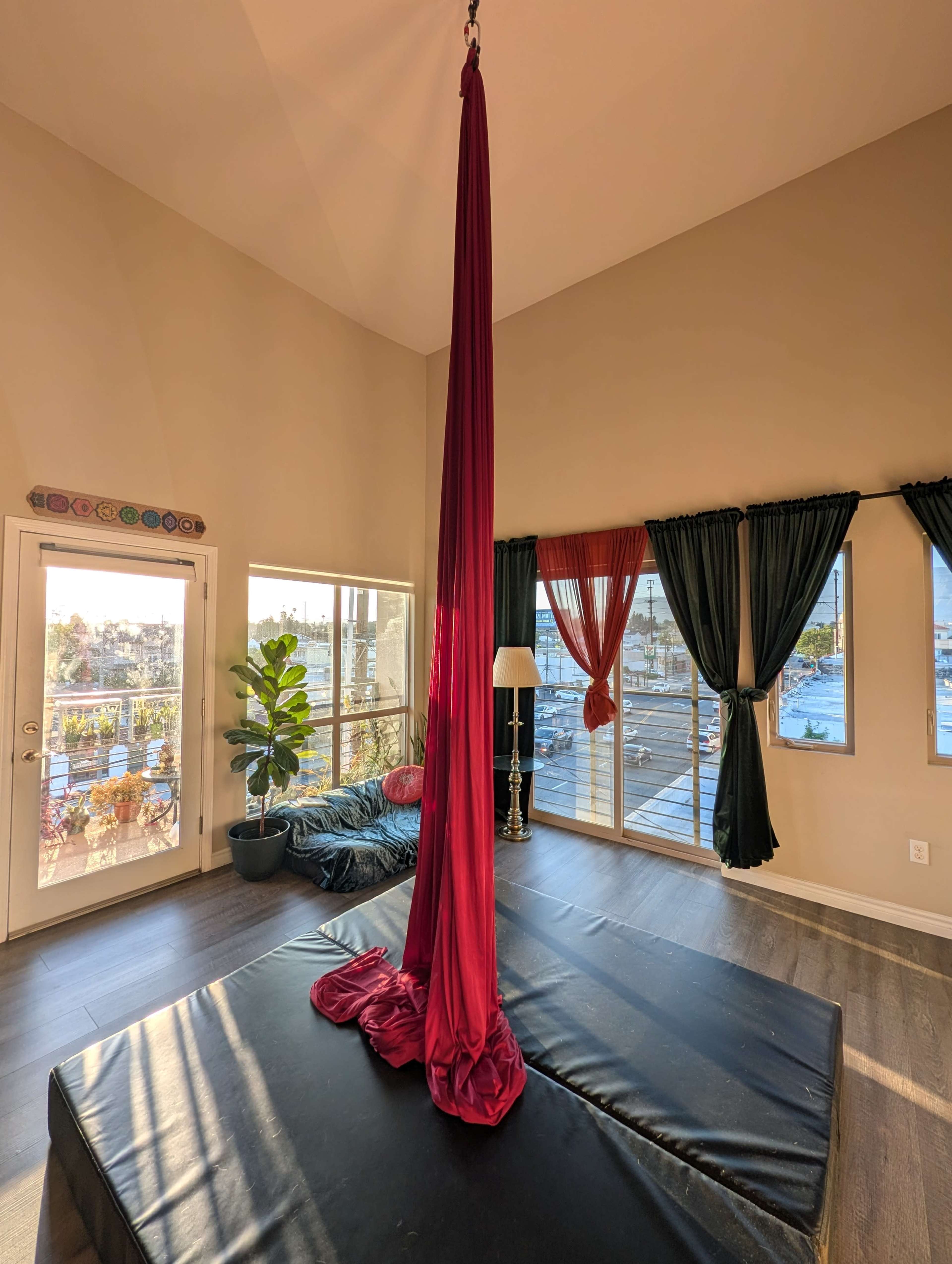 The image shows a spacious room with a tall red aerial silks hanging from the ceiling, surrounded by large windows and stylish curtains.