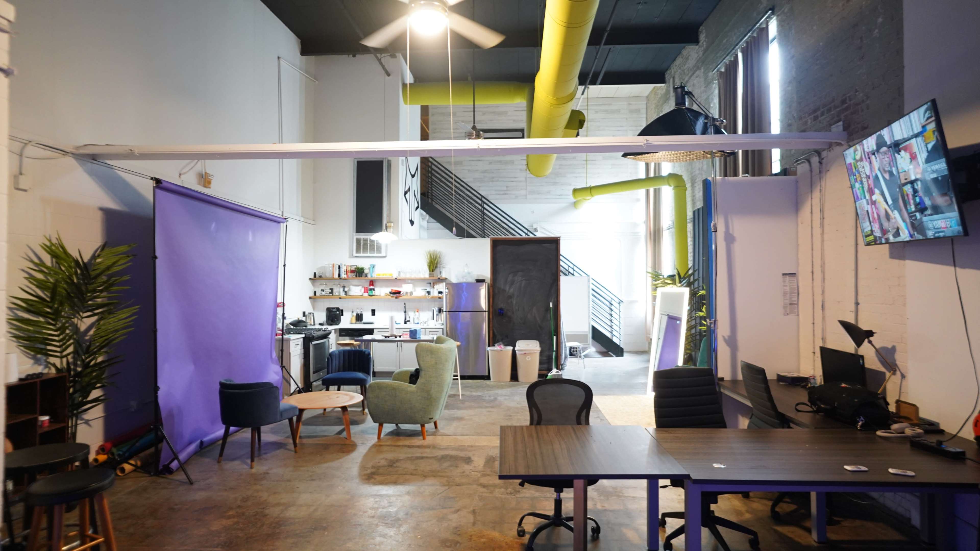 A modern office space features seating areas, a kitchen, and a staircase, with yellow ductwork adding color to the industrial aesthetic.